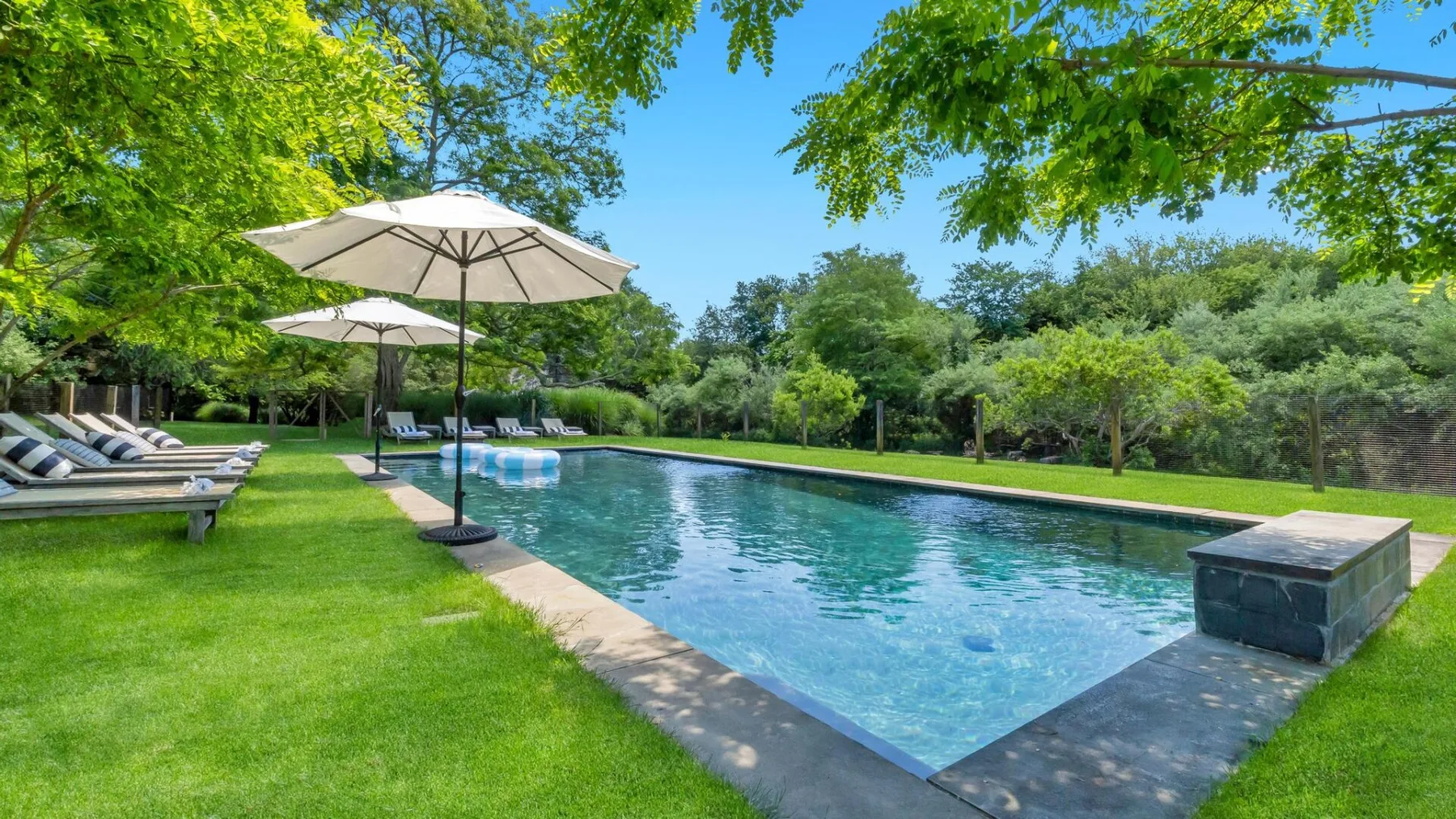 A rectangular swimming pool is surrounded by green lawn, trees, and patio umbrellas.