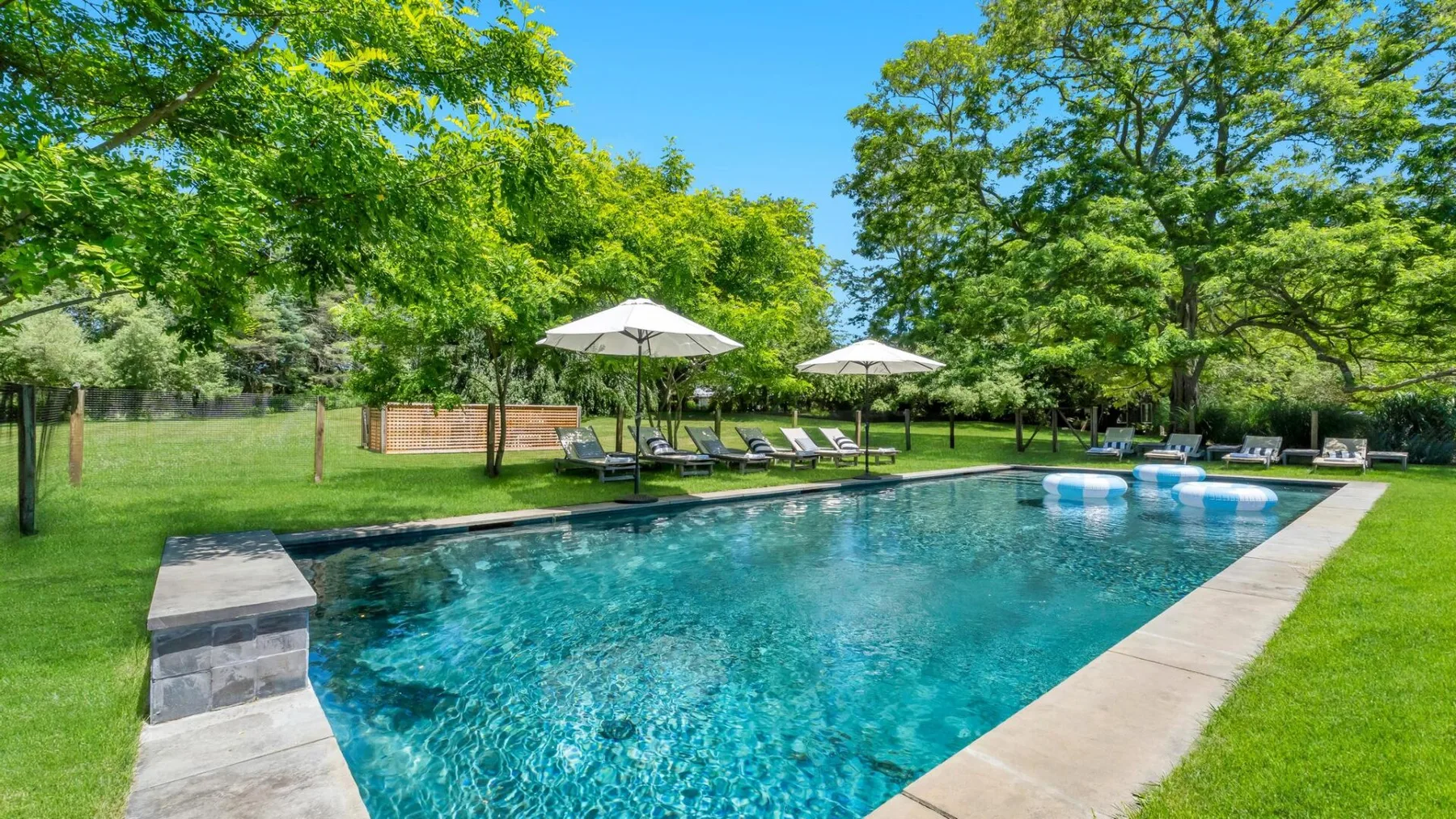 An outdoor swimming pool is surrounded by green lawns, trees, and shaded seating.