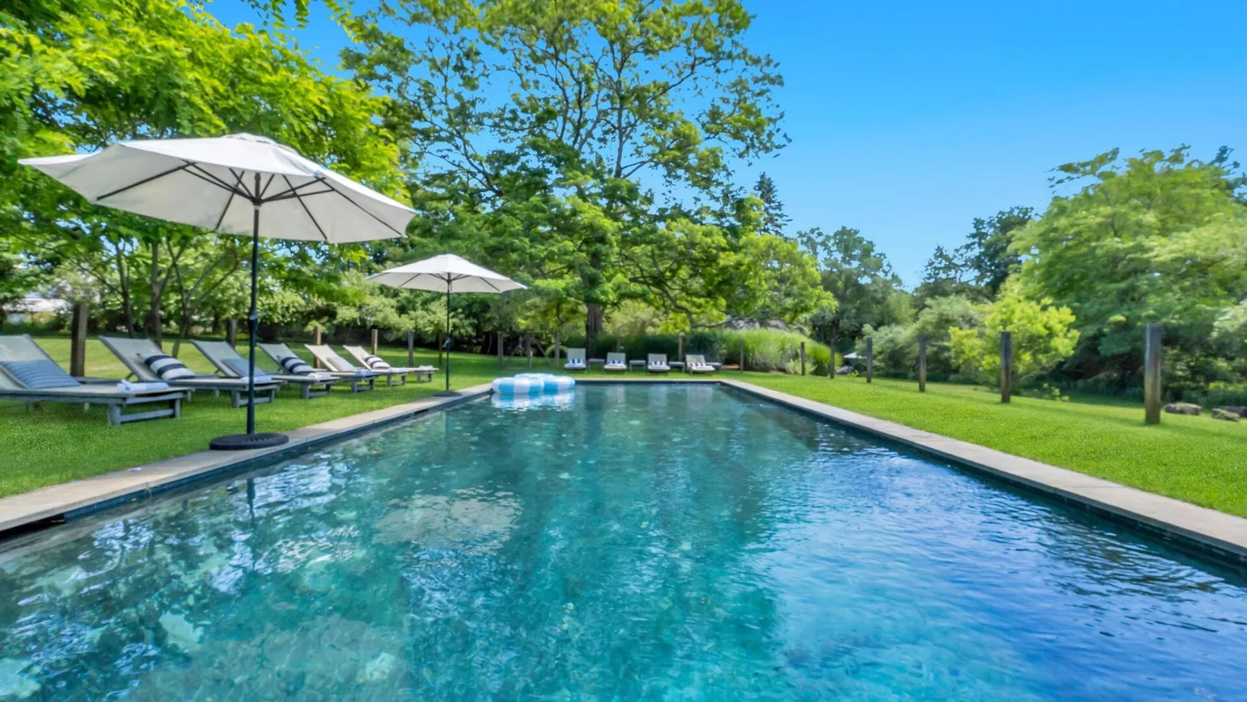A swimming pool is surrounded by green lawn, trees, and shaded seating.