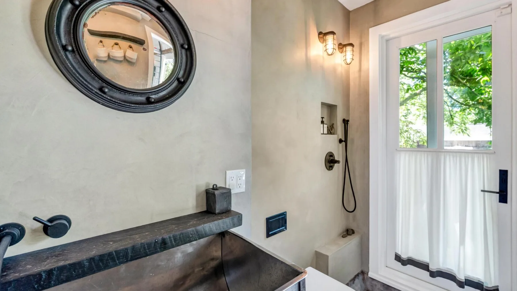 A bathroom with a dark vanity counter, wall mirror, and shower beside a bright window.
