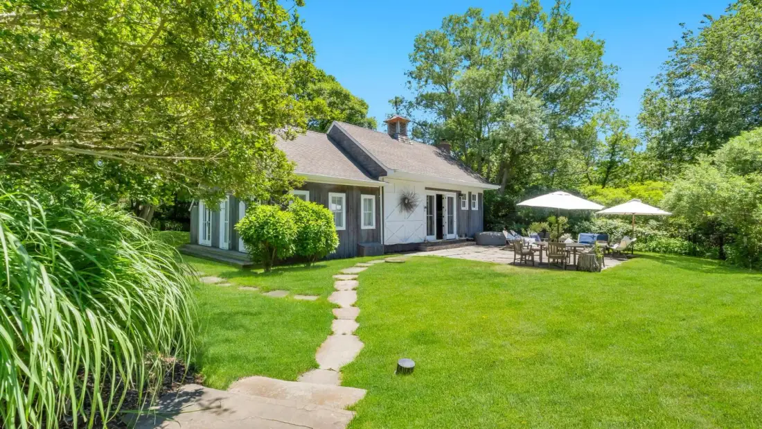 A small house stands beside a green lawn, with a stone path and outdoor seating.