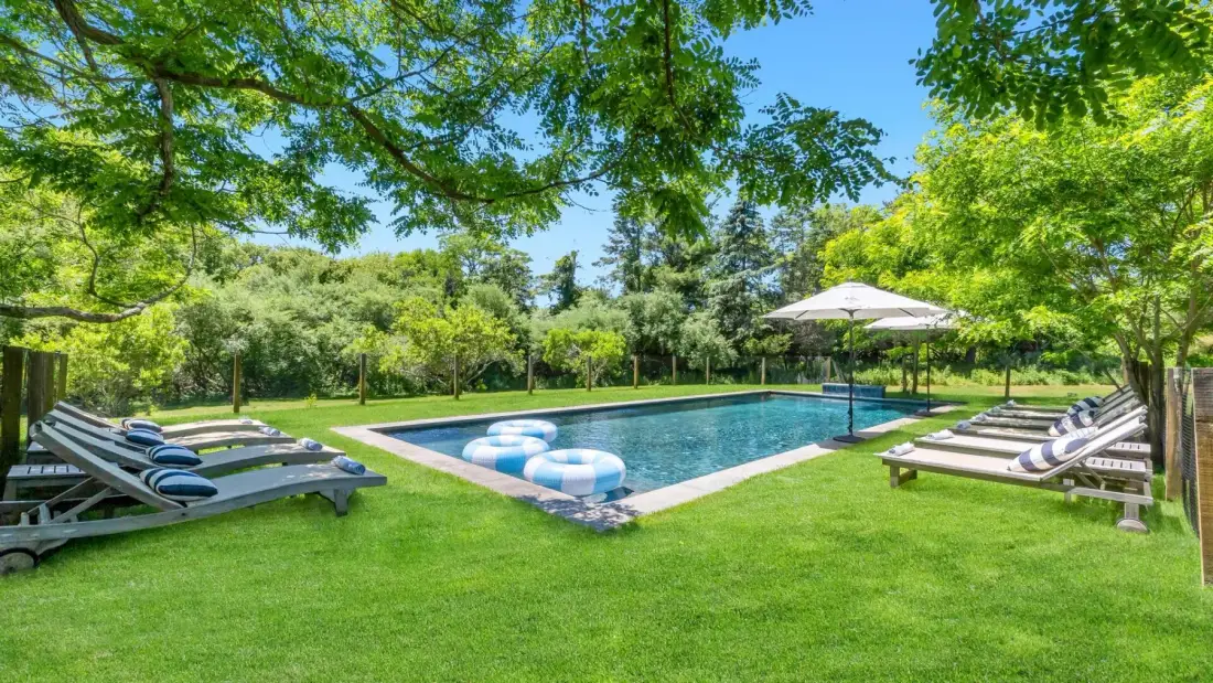 Una piscina está en un jardín verde con asientos reclinables y una sombrilla.