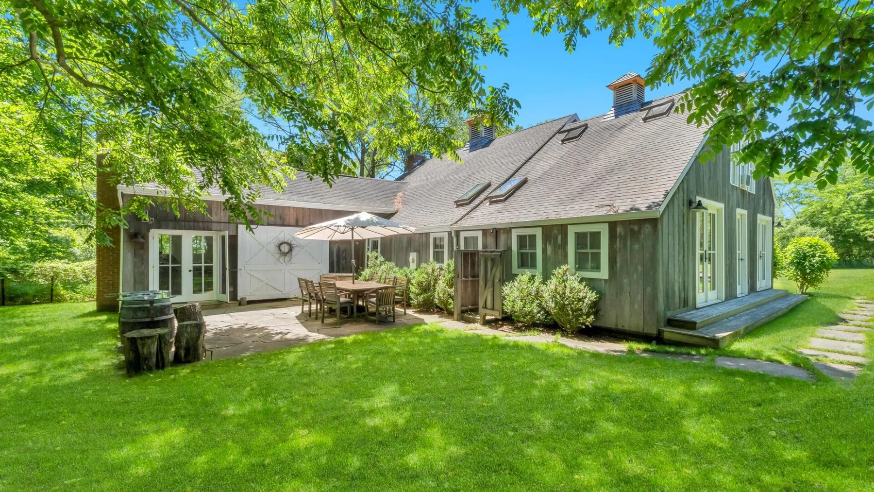 A house with a paved patio and outdoor dining area sits amid a green lawn and trees.