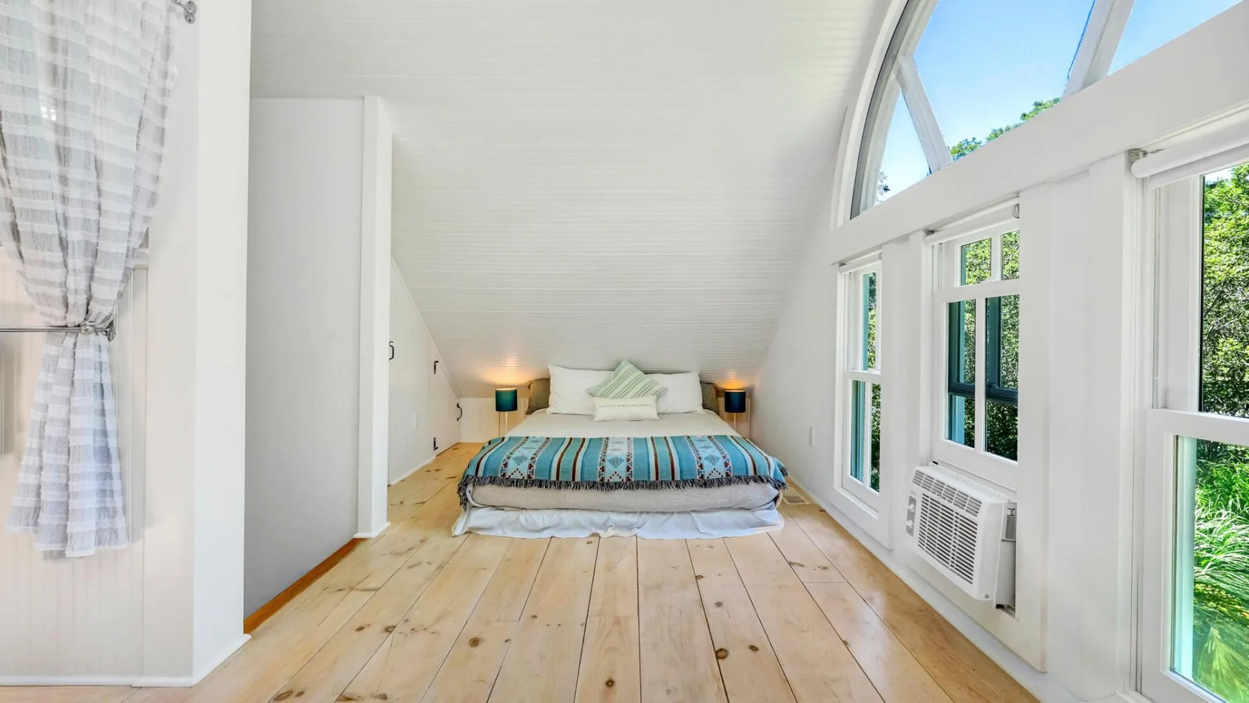 A bright bedroom with a bed beneath a sloped ceiling and large windows.