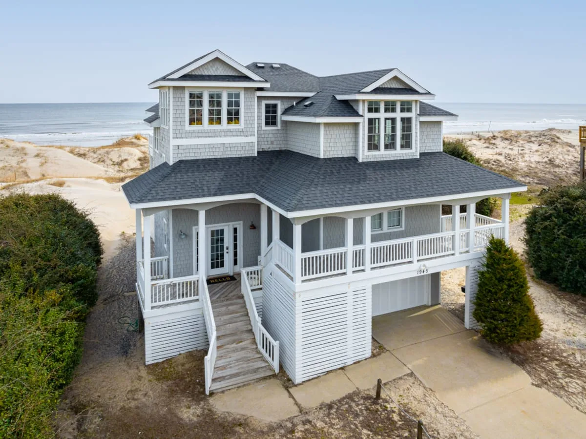 A coastal house with white siding stands beside sandy dunes and the ocean.