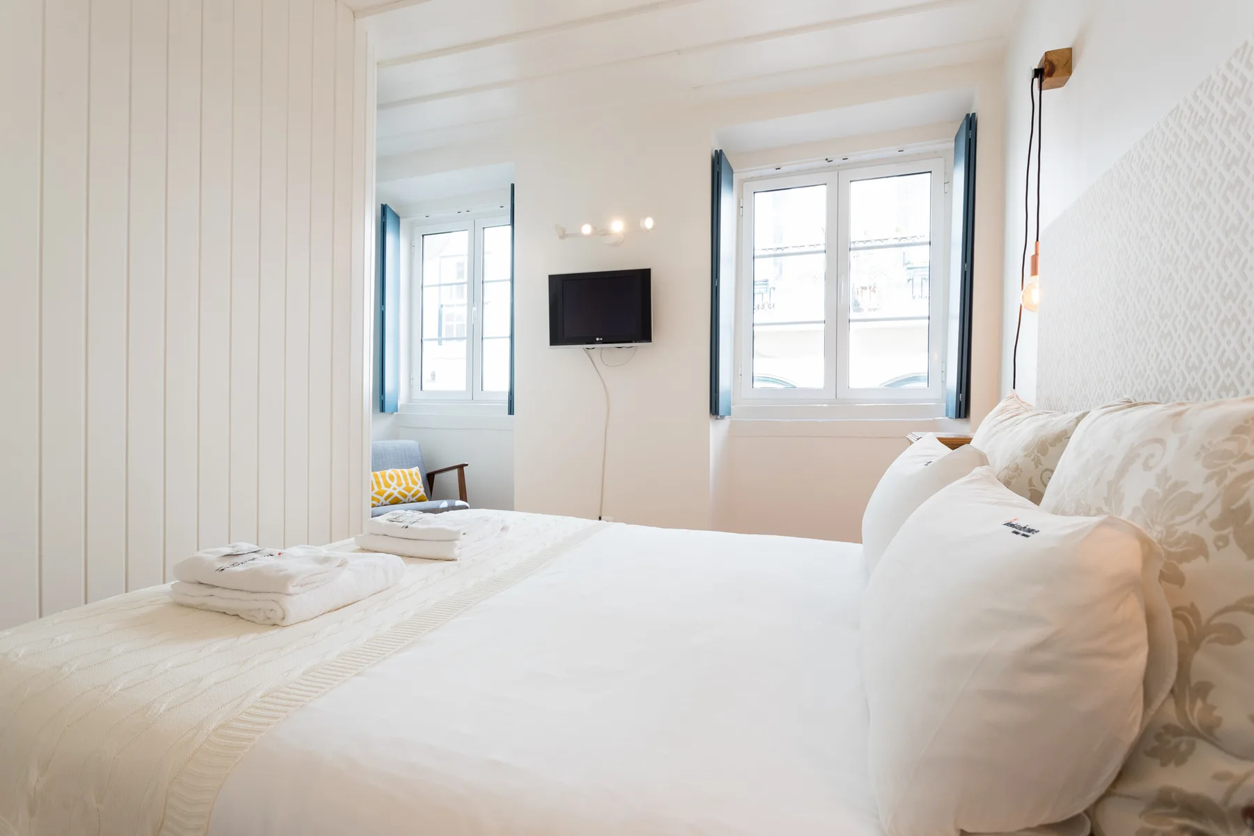 Un dormitorio luminoso con ropa de cama blanca y ventanas con vistas a la calle.