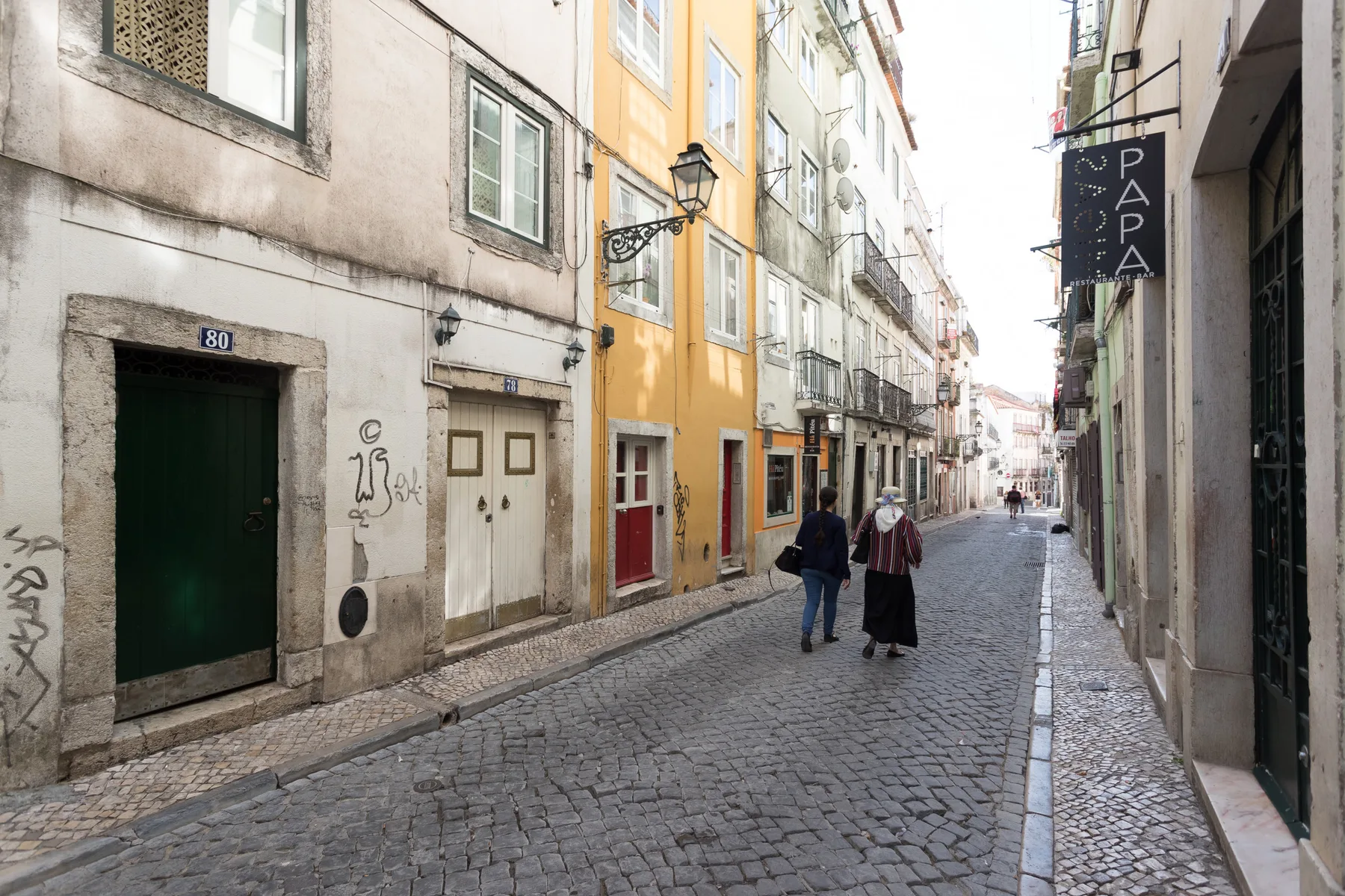 Una calle adoquinada pasa entre edificios cercanos, con dos peatones caminando por ella.