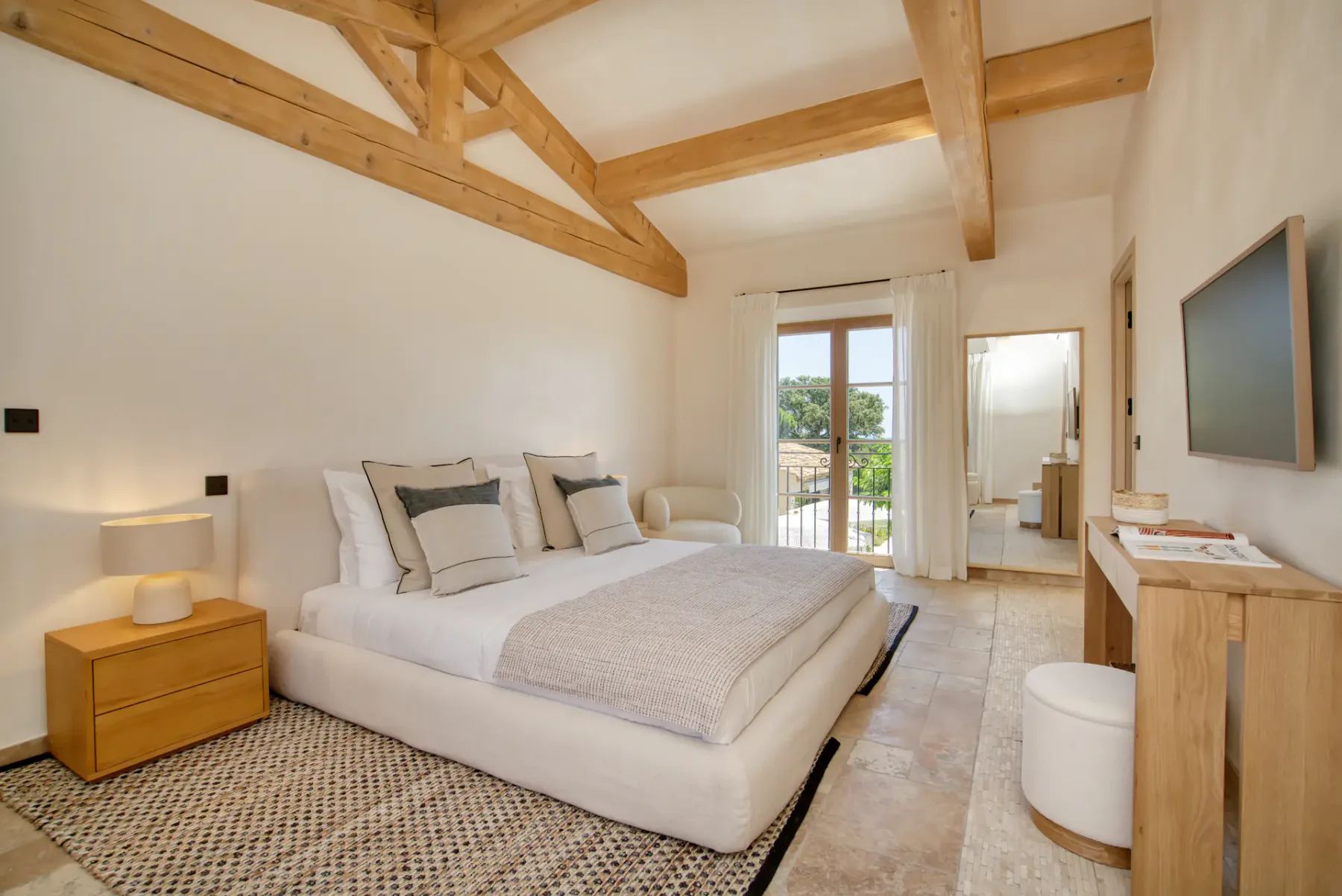 Un dormitorio acogedor con una cama grande, vigas de madera, una mesita de noche, un televisor montado en la pared, un espejo y una ventana con cortinas con vistas a la vegetación.
