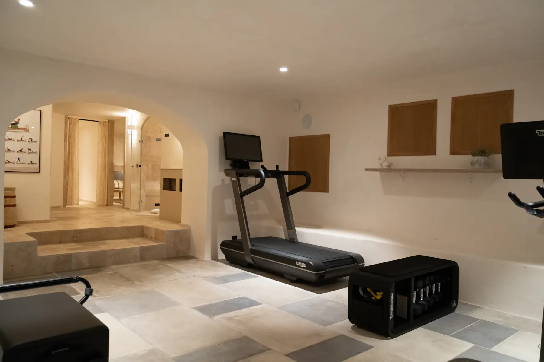 Gimnasio moderno en casa con una cinta de correr, equipos de ejercicio y suelo de baldosas beige. Un arco conduce a una habitación iluminada al fondo.