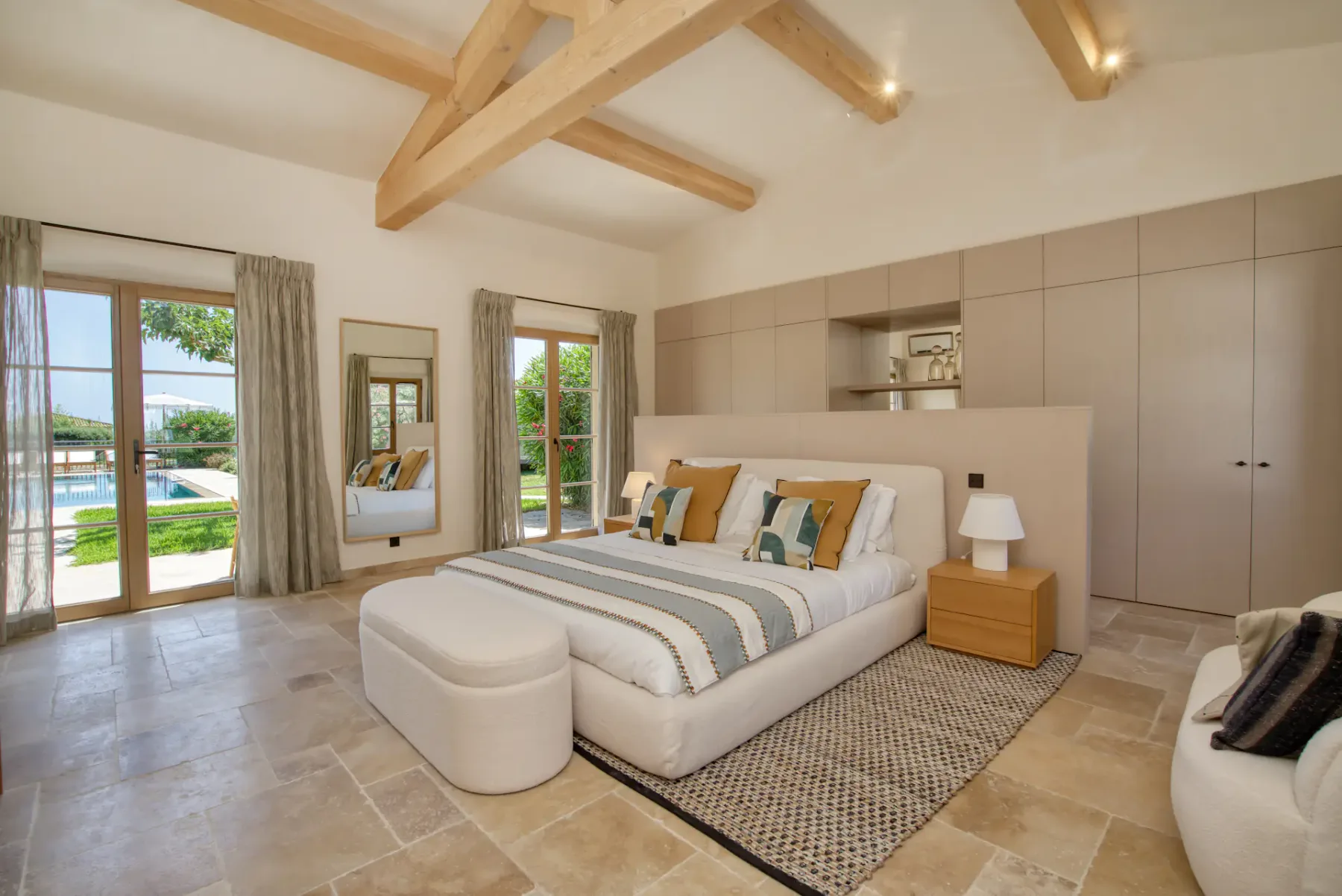 Dormitorio amplio con vigas de madera, cama grande, puf y ventanas con vistas a una piscina y un jardín. Tonos neutros y decoración moderna.