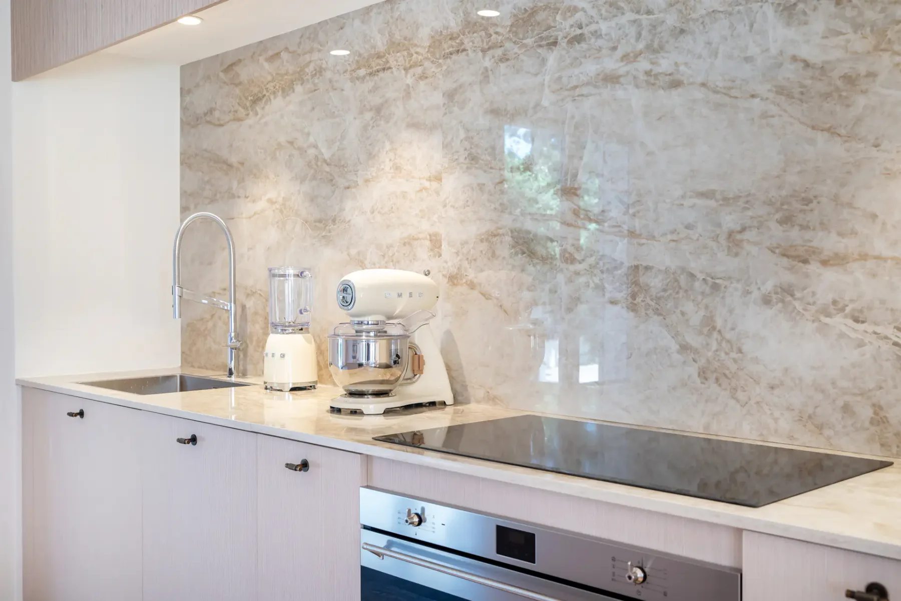 Cocina moderna con revestimiento de mármol en la pared de la encimera, horno empotrado, placa de cocción y pequeños electrodomésticos sobre la encimera, entre ellos una batidora de vaso y una batidora de pedestal.