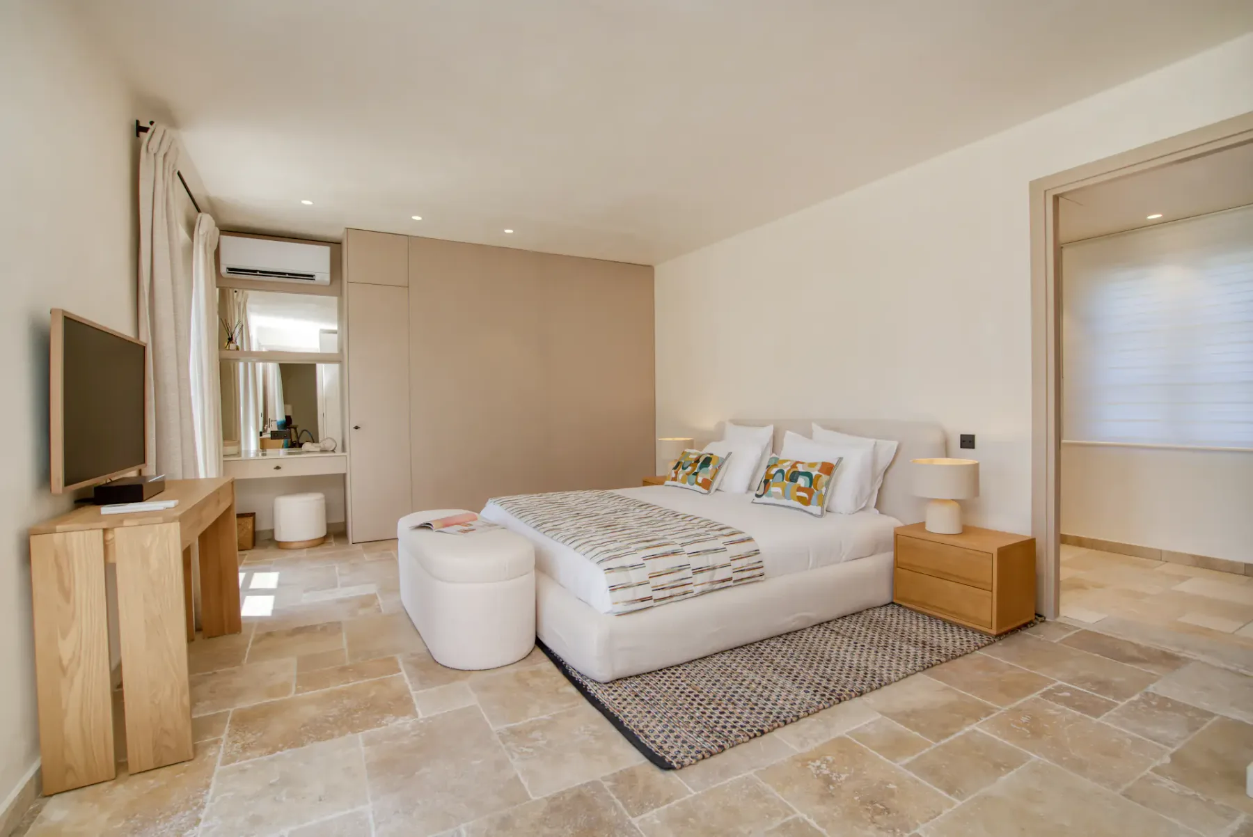 Dormitorio moderno con cama doble, cojines decorativos, mesitas de noche, un televisor montado en la pared, un tocador y suelo de baldosas beige.