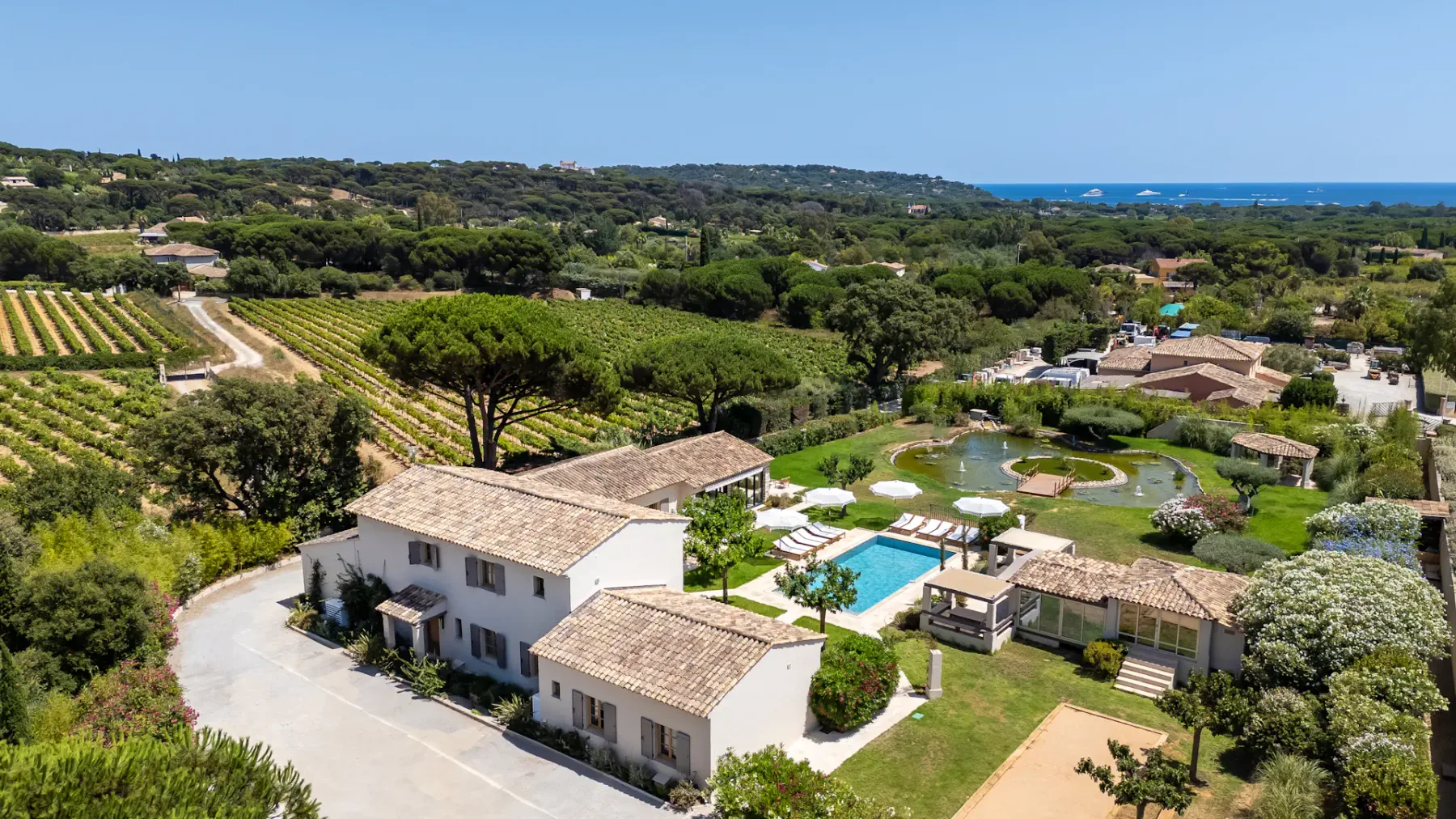 Vista aérea de una villa rodeada de viñedos, una piscina y un paisaje exuberante, con el mar visible a lo lejos bajo un cielo azul despejado.