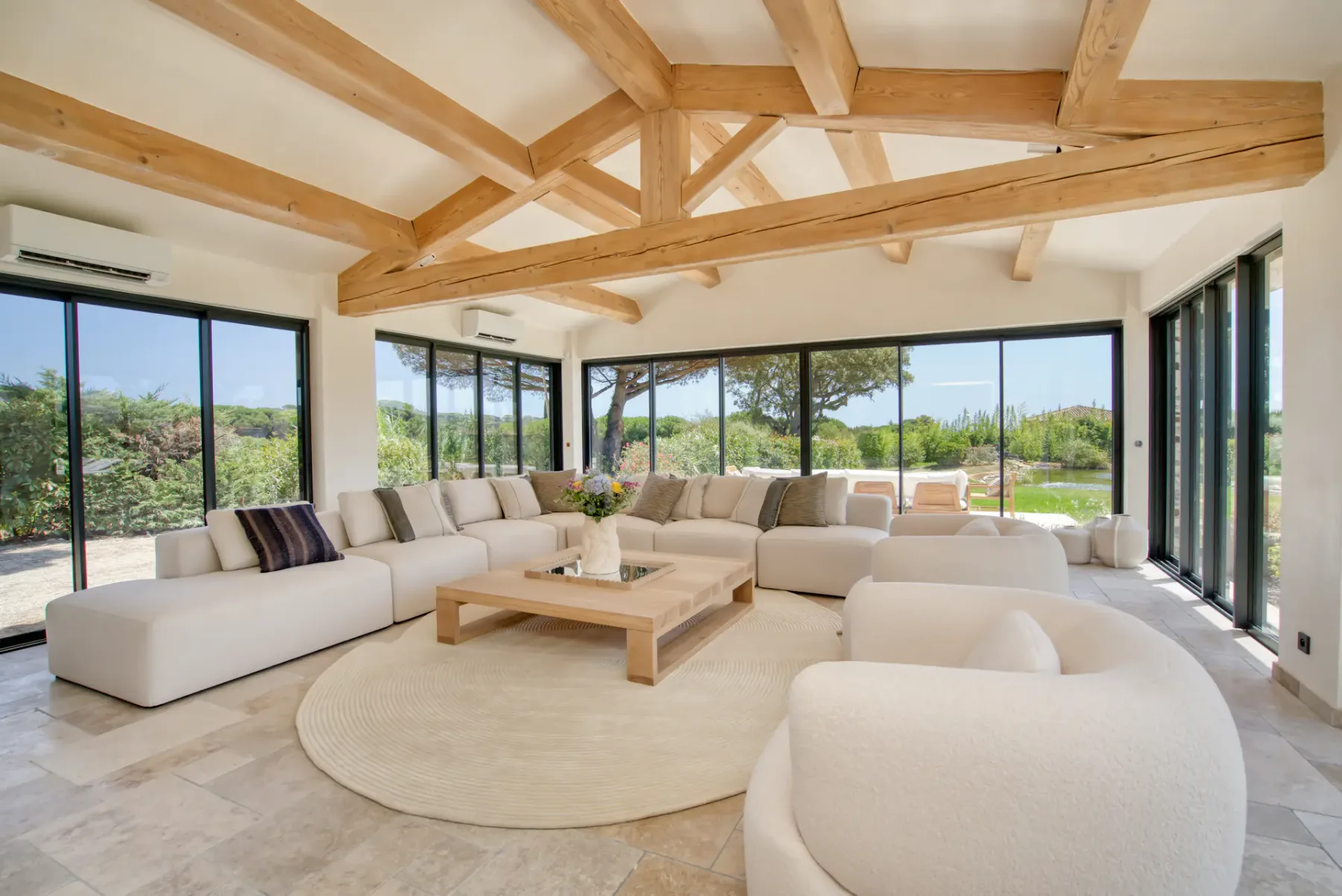 Amplio salón con vigas de madera vistas, un gran sofá modular, sillones redondos, mesa de centro y ventanales de suelo a techo con vistas a la vegetación.