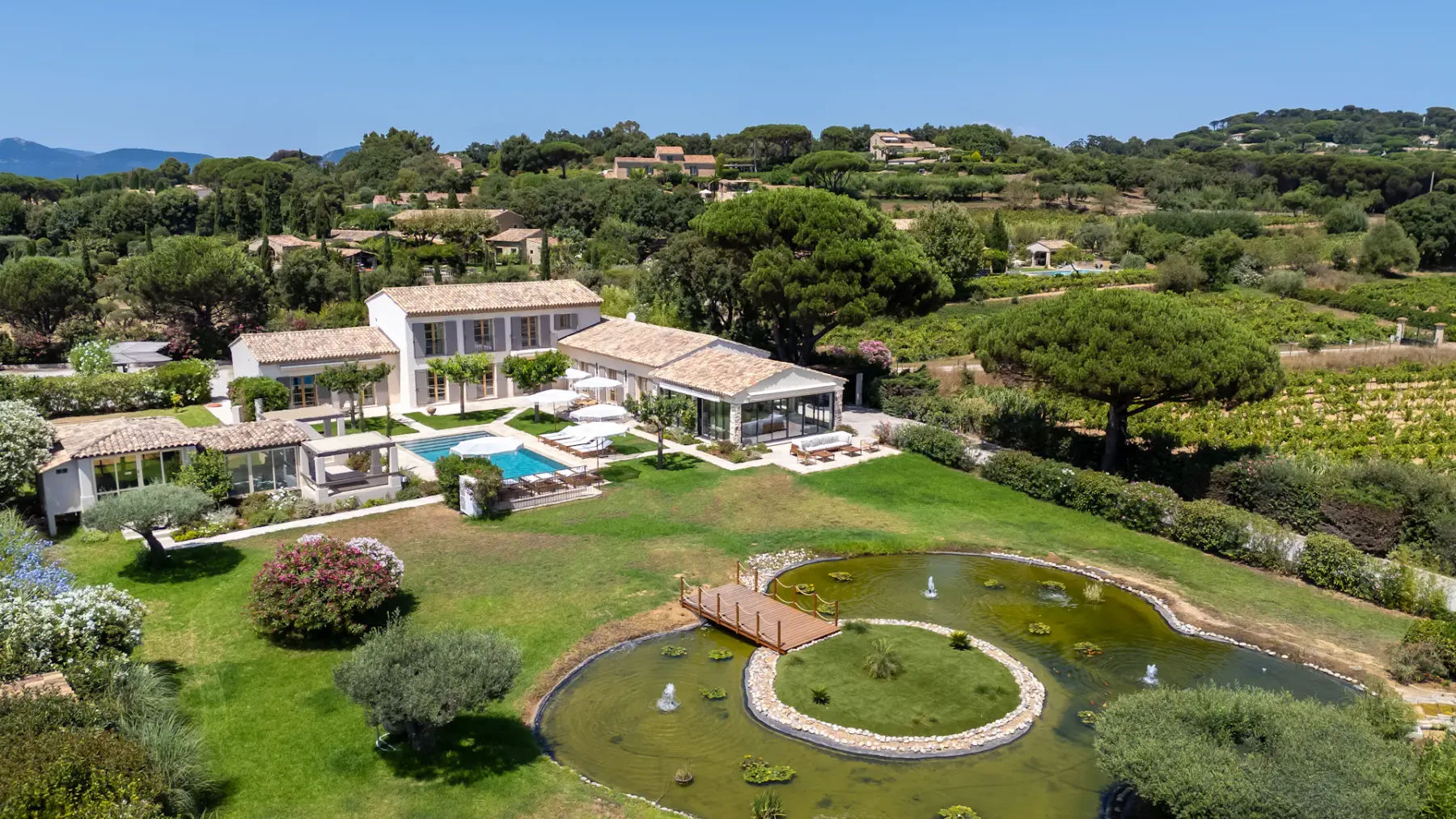 Vista aérea de una lujosa villa con piscina, jardines exuberantes y un estanque, situada en un pintoresco paisaje rural bajo un cielo azul despejado.