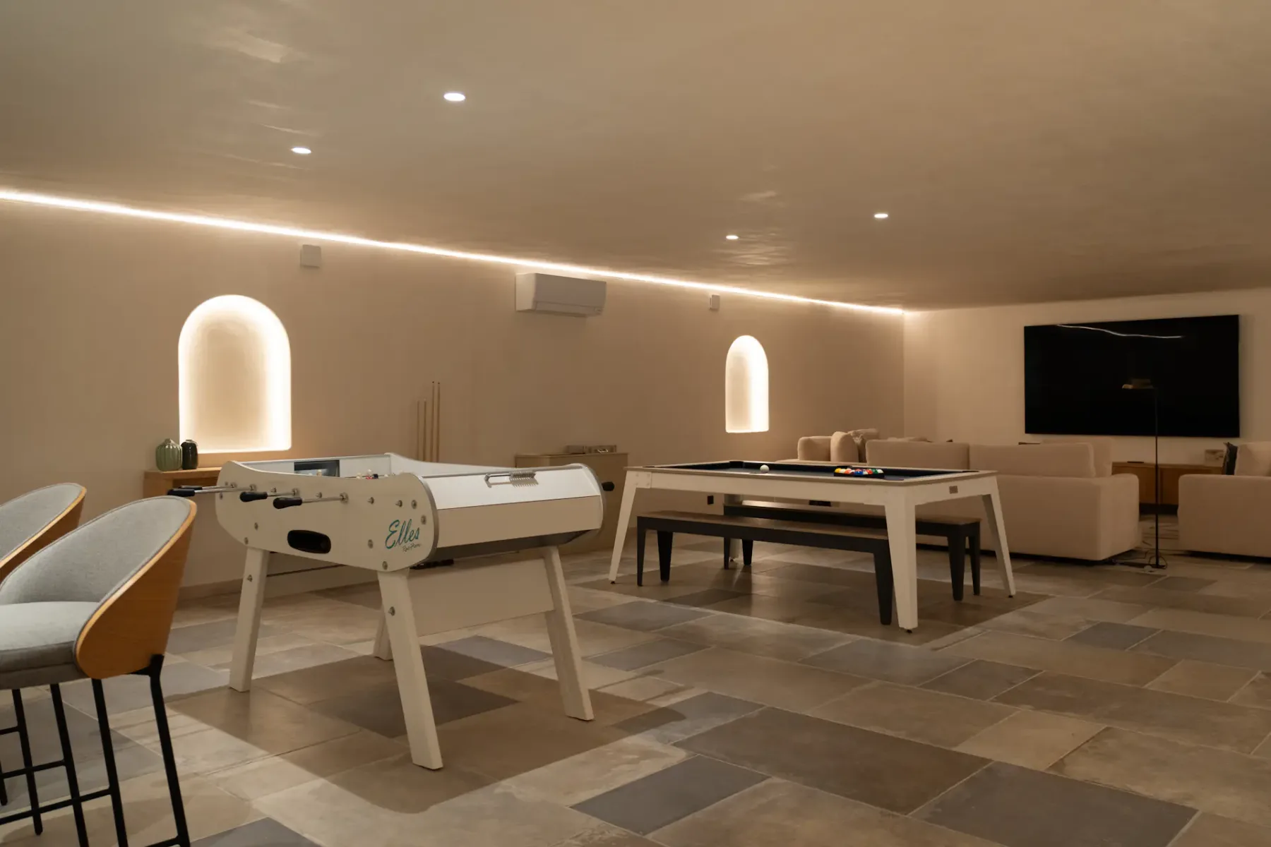 Sala amplia con un futbolín, una mesa de billar, taburetes de bar y un televisor grande. La iluminación moderna y los tonos neutros crean un ambiente acogedor.