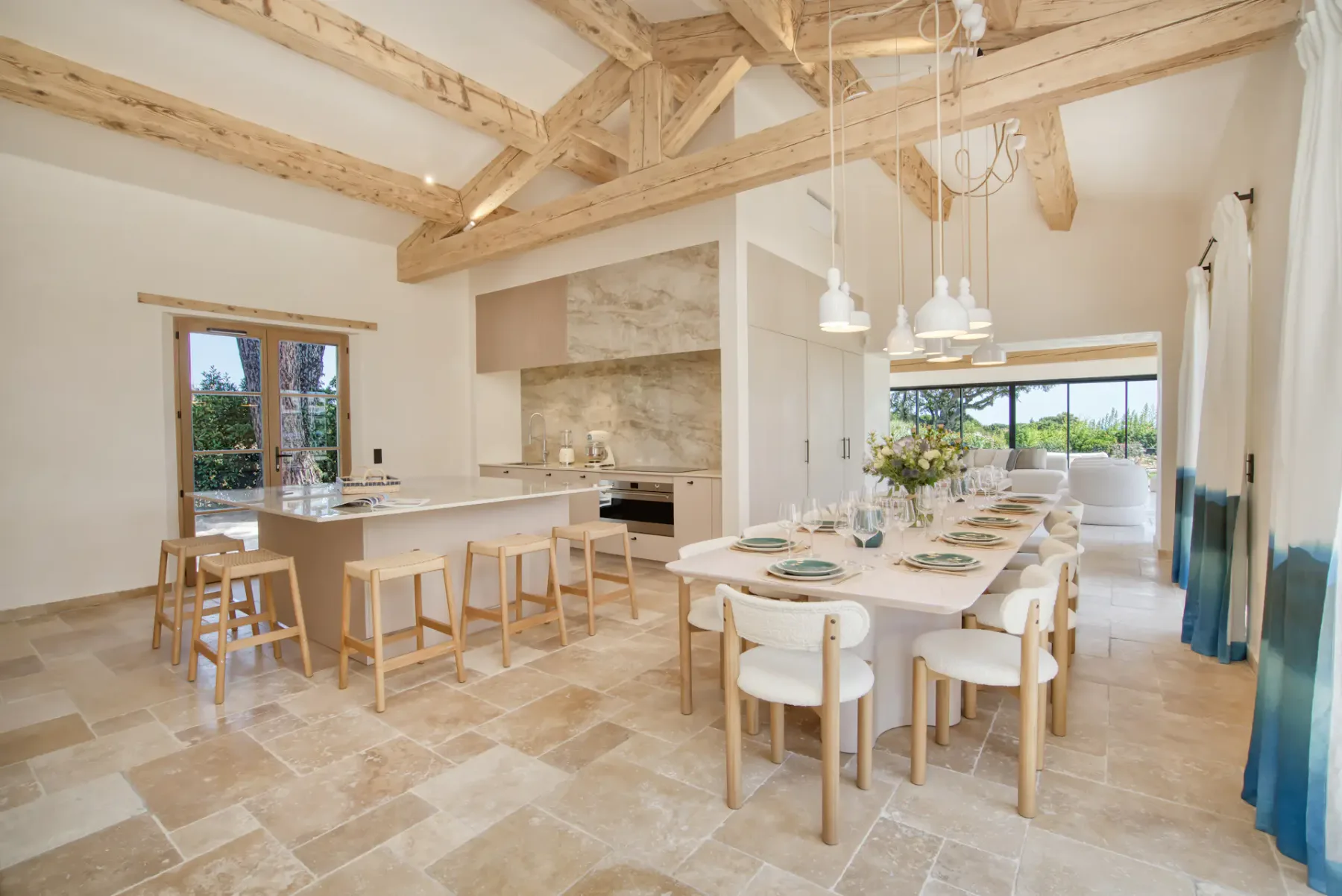 Amplia cocina y zona de comedor con vigas de madera, suelo de piedra, electrodomésticos modernos y una gran mesa puesta con platos y copas. La luz natural inunda el espacio.