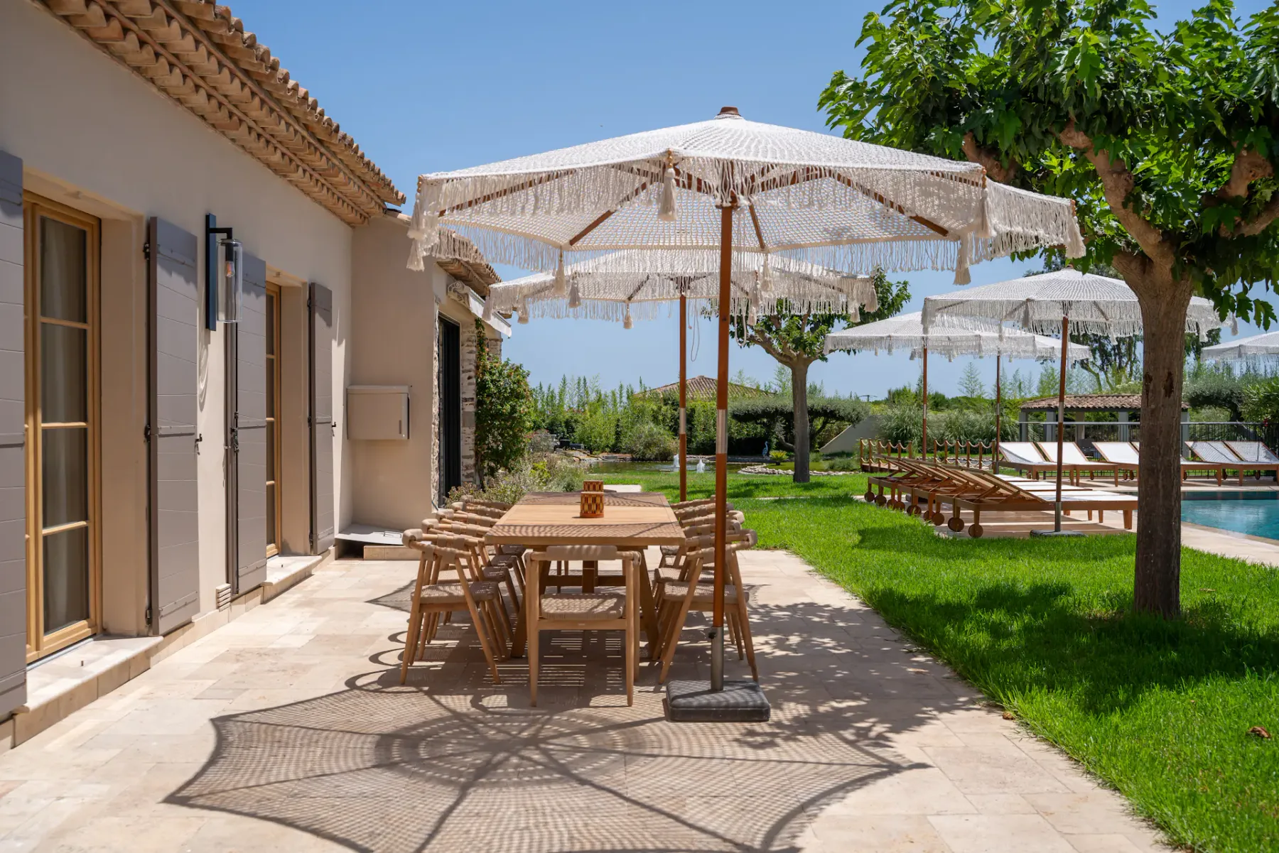 Un patio soleado con mesas de comedor al aire libre y sombrillas con flecos junto a un césped verde.