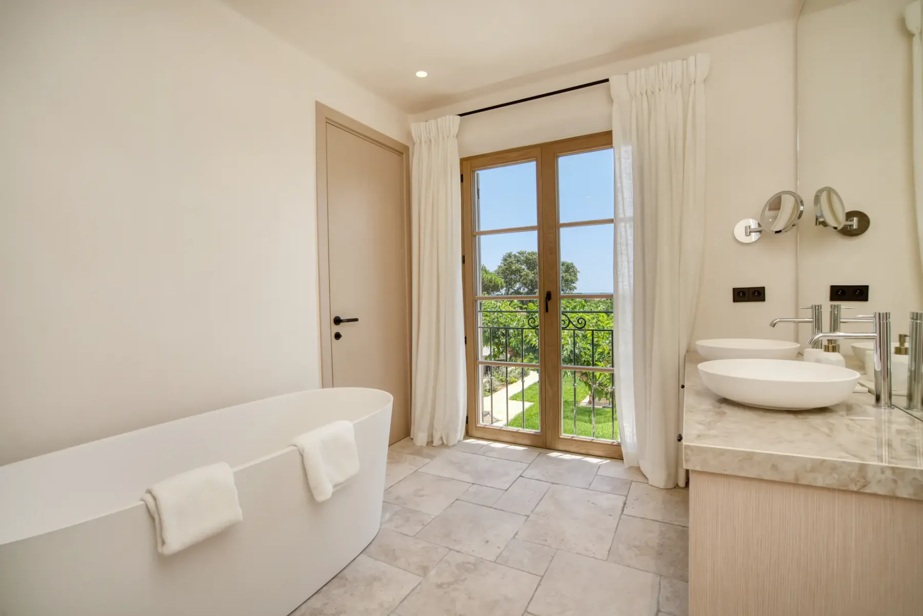 Baño lujoso con una bañera exenta, dos lavabos, una ventana grande con vistas al jardín y baldosas de tonos claros.