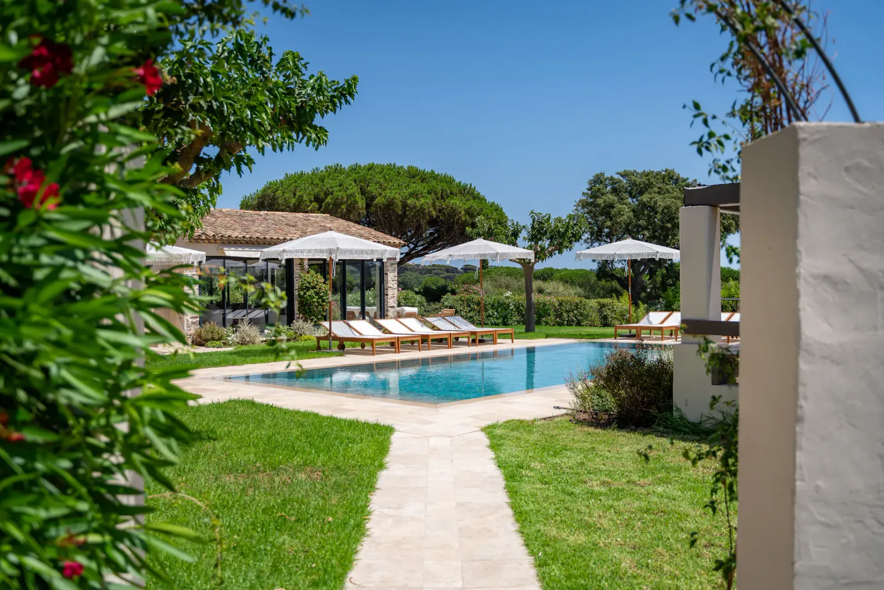 Una tranquila zona de piscina al aire libre con tumbonas y sombrillas, rodeada de vegetación exuberante y bajo un cielo azul despejado.