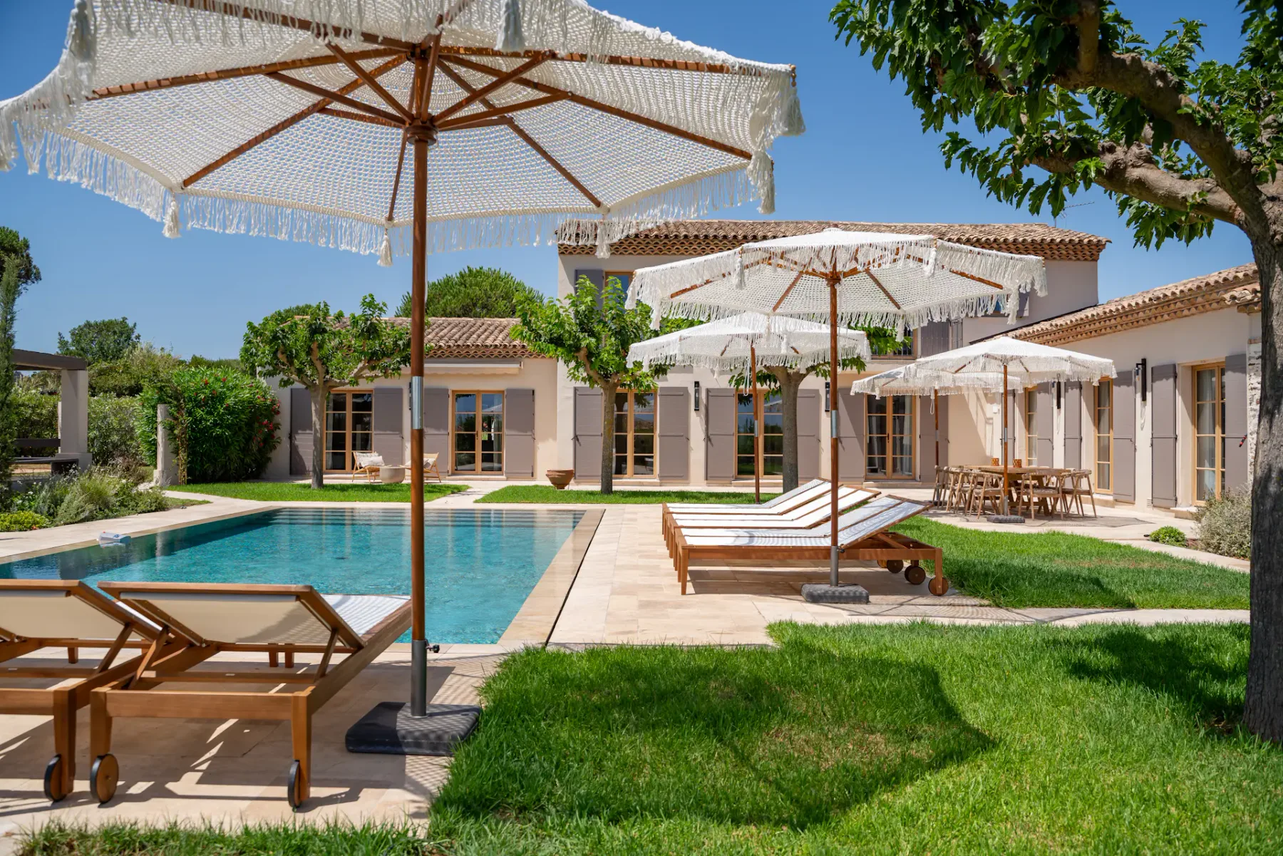 Lujosa villa con piscina, rodeada de tumbonas y grandes sombrillas, situada en un jardín exuberante bajo un cielo azul despejado.