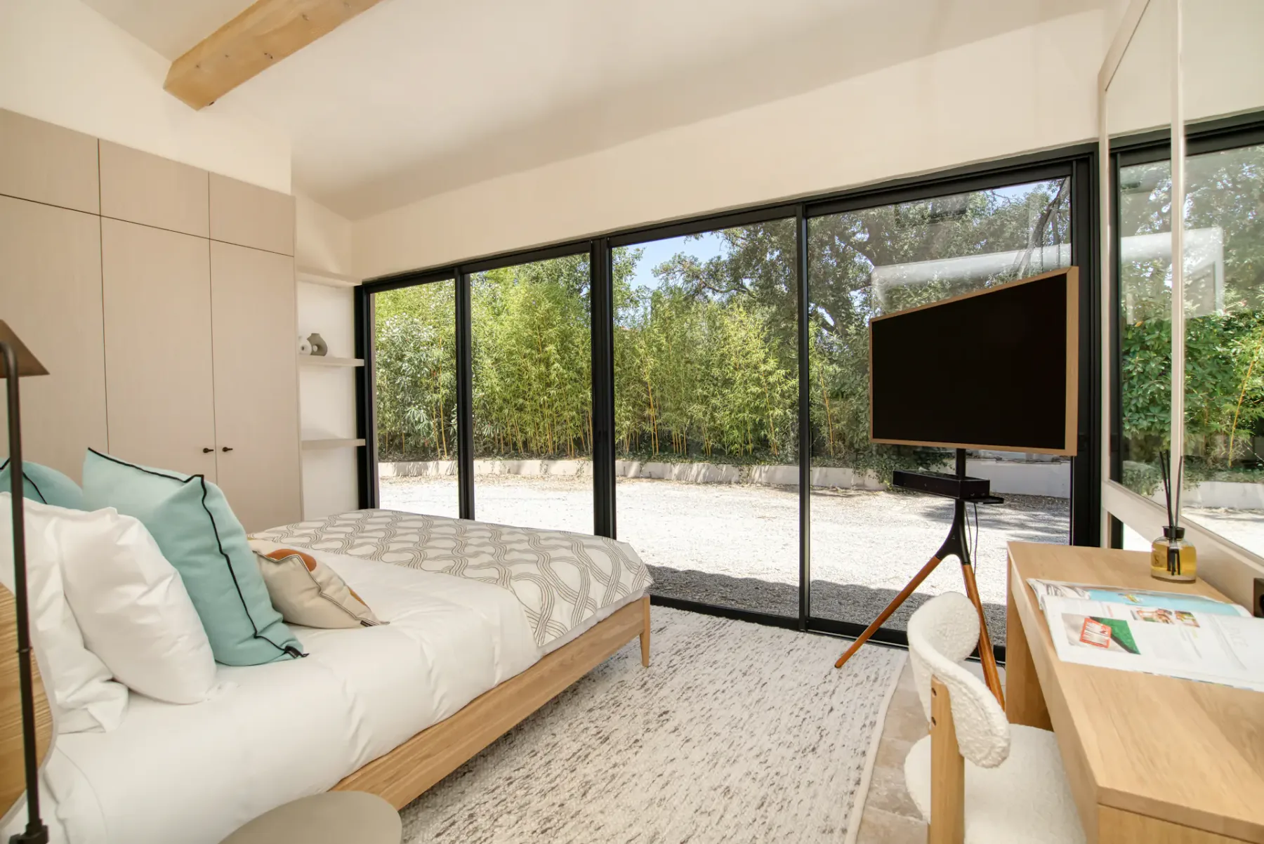 Dormitorio moderno con grandes ventanales, una cama bien hecha, muebles de madera y vistas a un jardín con árboles y vegetación en el exterior.