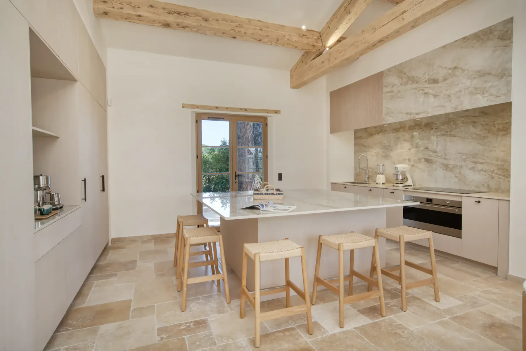 Cocina moderna con vigas de madera, suelo de baldosas de piedra, una isla central con taburetes de bar y grandes ventanales con vistas a la vegetación.