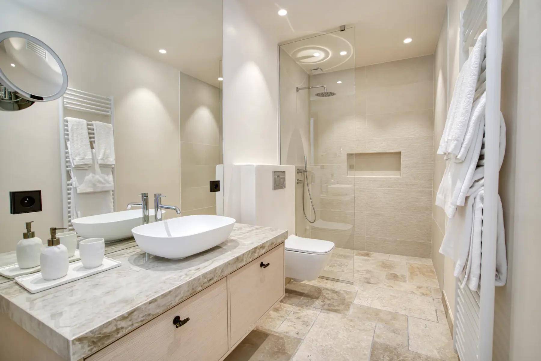 Baño moderno con encimera de mármol, lavabo sobre encimera, grifo de pared, ducha a ras de suelo y toallero. Paleta de tonos neutros e iluminación empotrada.