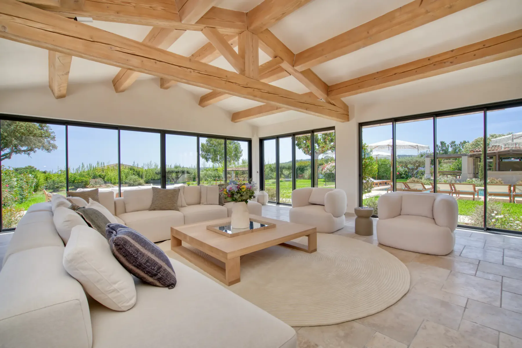 Amplio salón con un gran sofá modular beige, mesa de centro de madera y techo con vigas vistas, con vistas a un jardín exuberante a través de puertas de cristal.