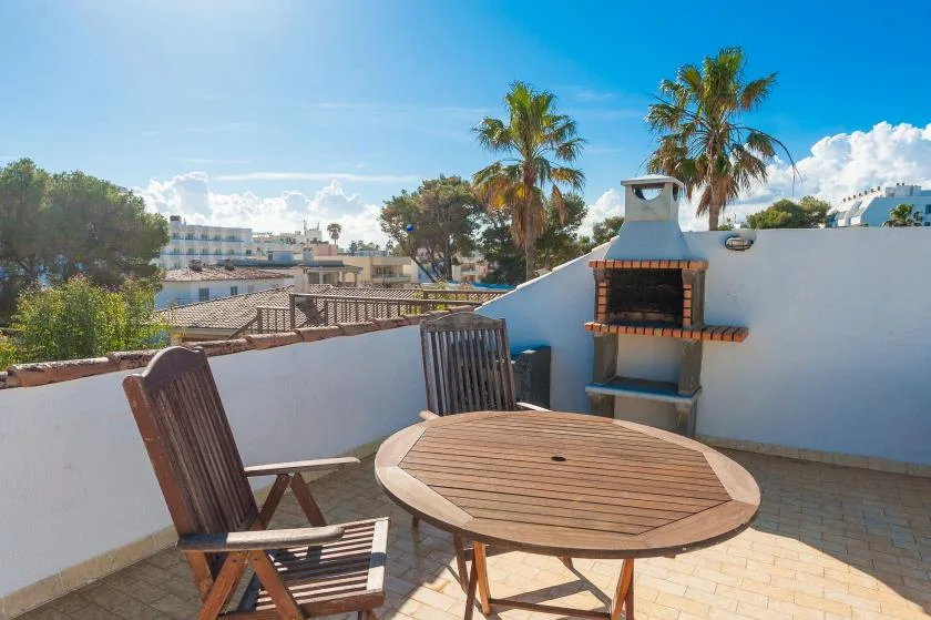 Una terraza soleada con una mesa redonda de madera, asientos y una barbacoa empotrada.