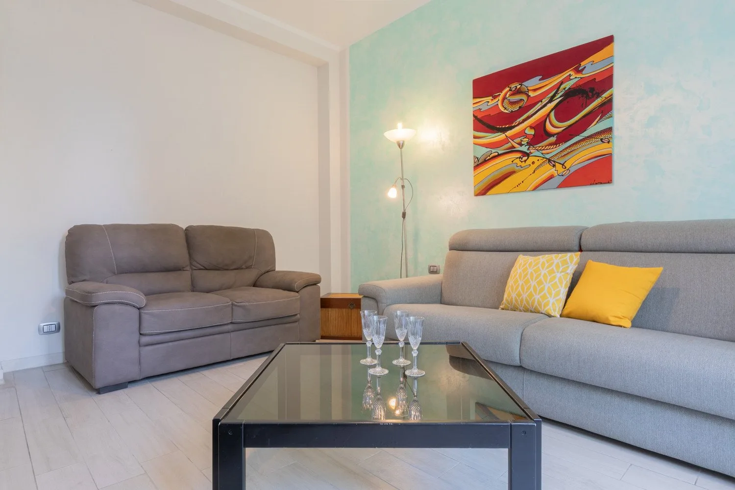 Una sala de estar con asientos grises, una mesa de centro de vidrio y arte colorido en la pared.
