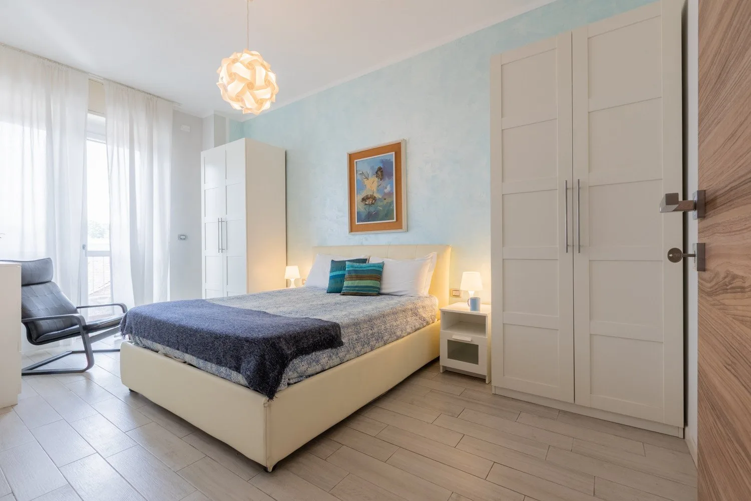 Un dormitorio con una cama doble, mesitas de noche y una silla junto a la ventana.