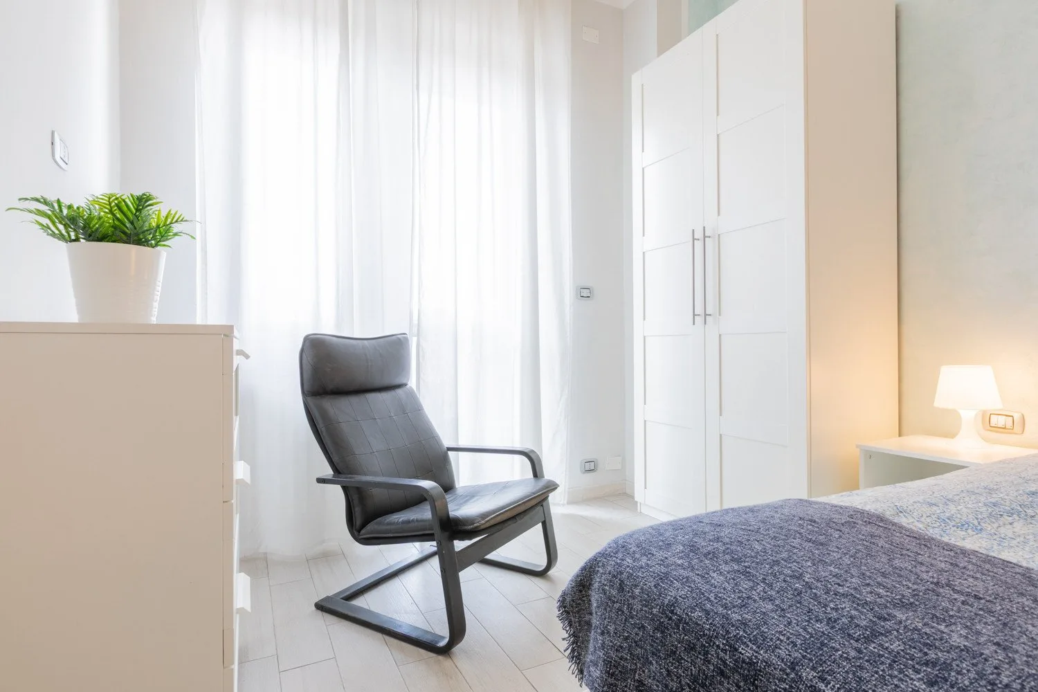 Un dormitorio luminoso con un sillón oscuro junto a la cama y un armario alto.