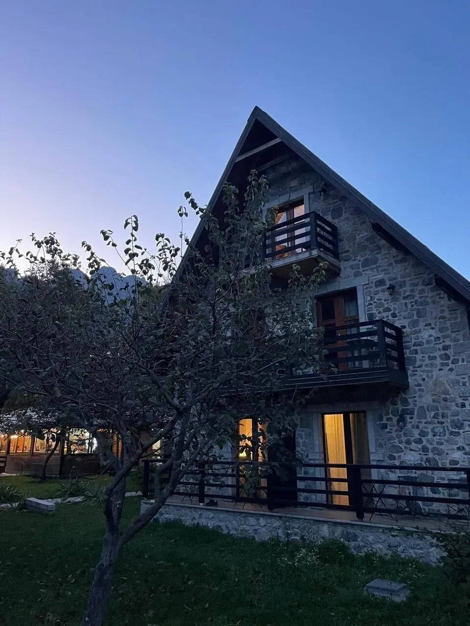 Edificio de piedra del Gjeçaj Guesthouse al anochecer, con ventanas iluminadas con luz cálida.