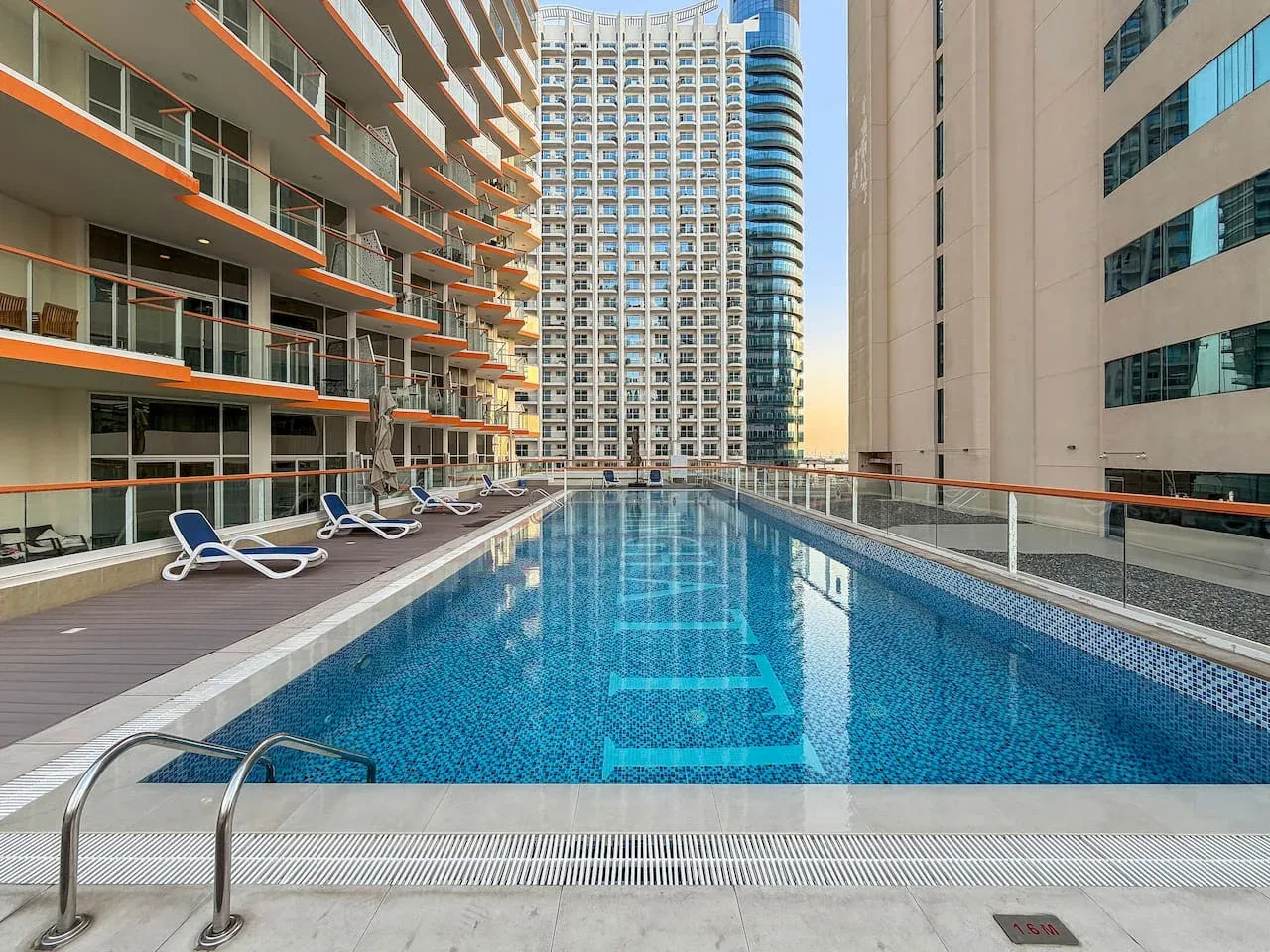 Una piscina al aire libre está entre edificios altos, con tumbonas junto a la terraza.