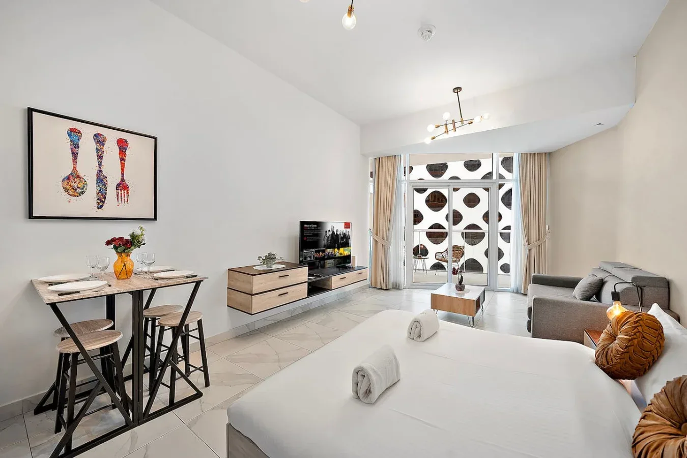 Una habitación luminosa combina cama, mesa de comedor, televisor y asientos junto a ventanas estampadas.