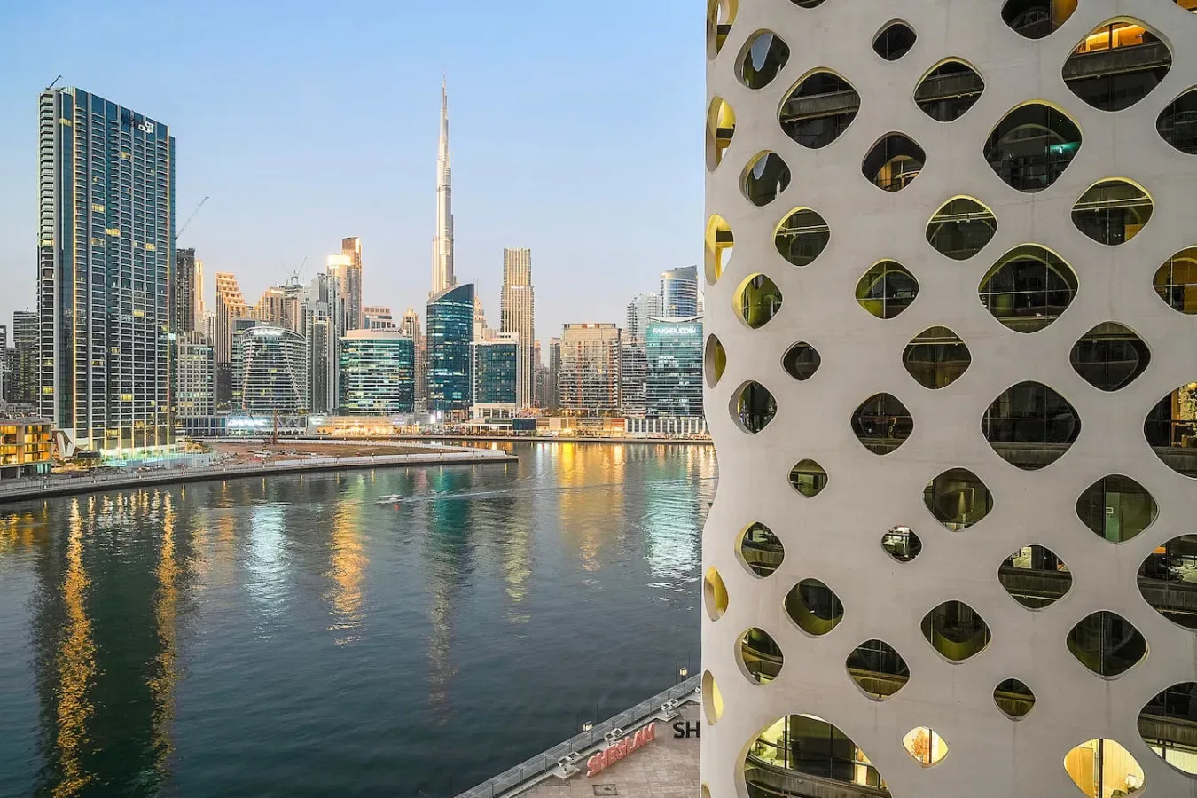 A waterfront city skyline reflects across the water beside a patterned building facade.