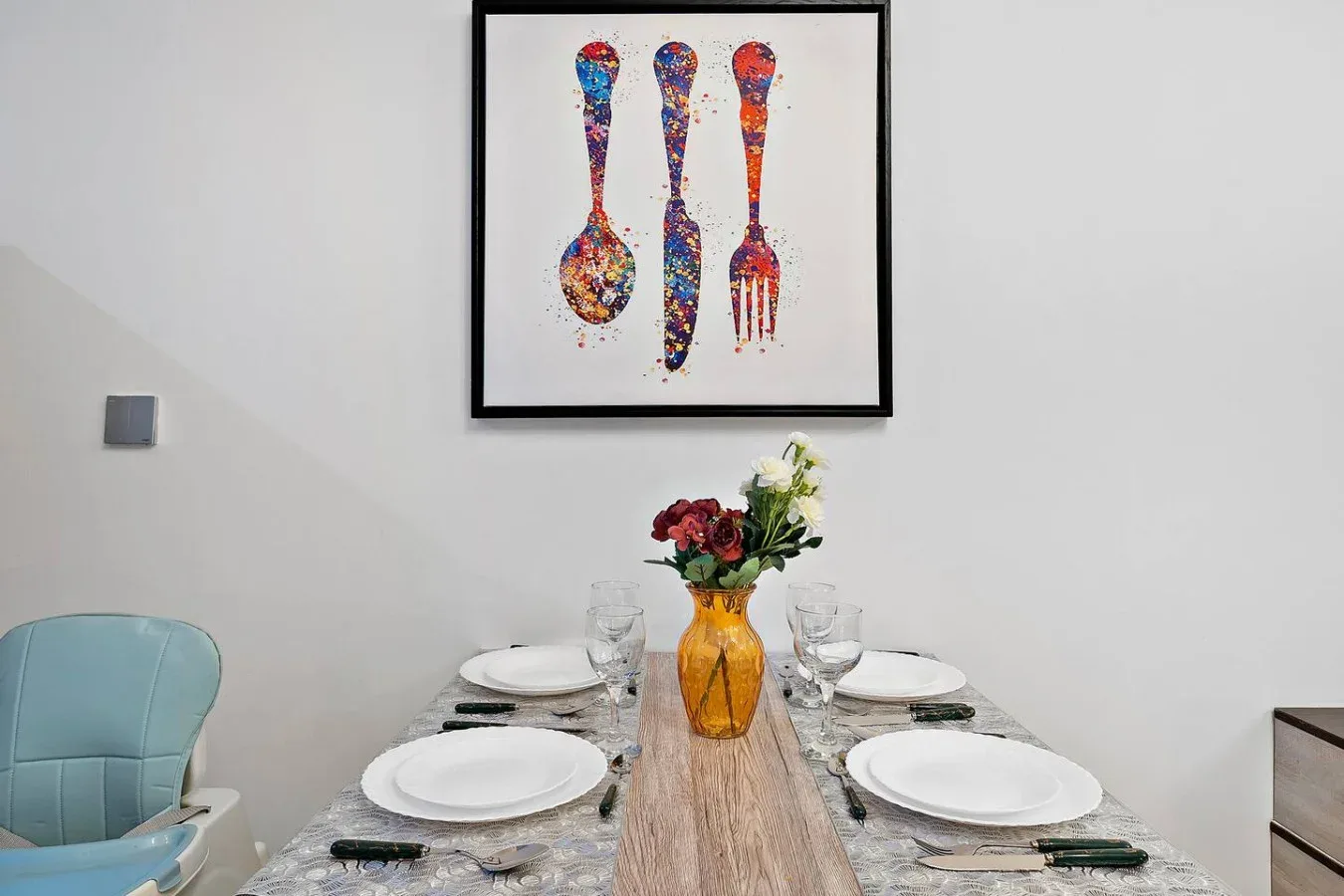 Una mesa de comedor está puesta con platos y un jarrón de flores bajo un cuadro enmarcado.