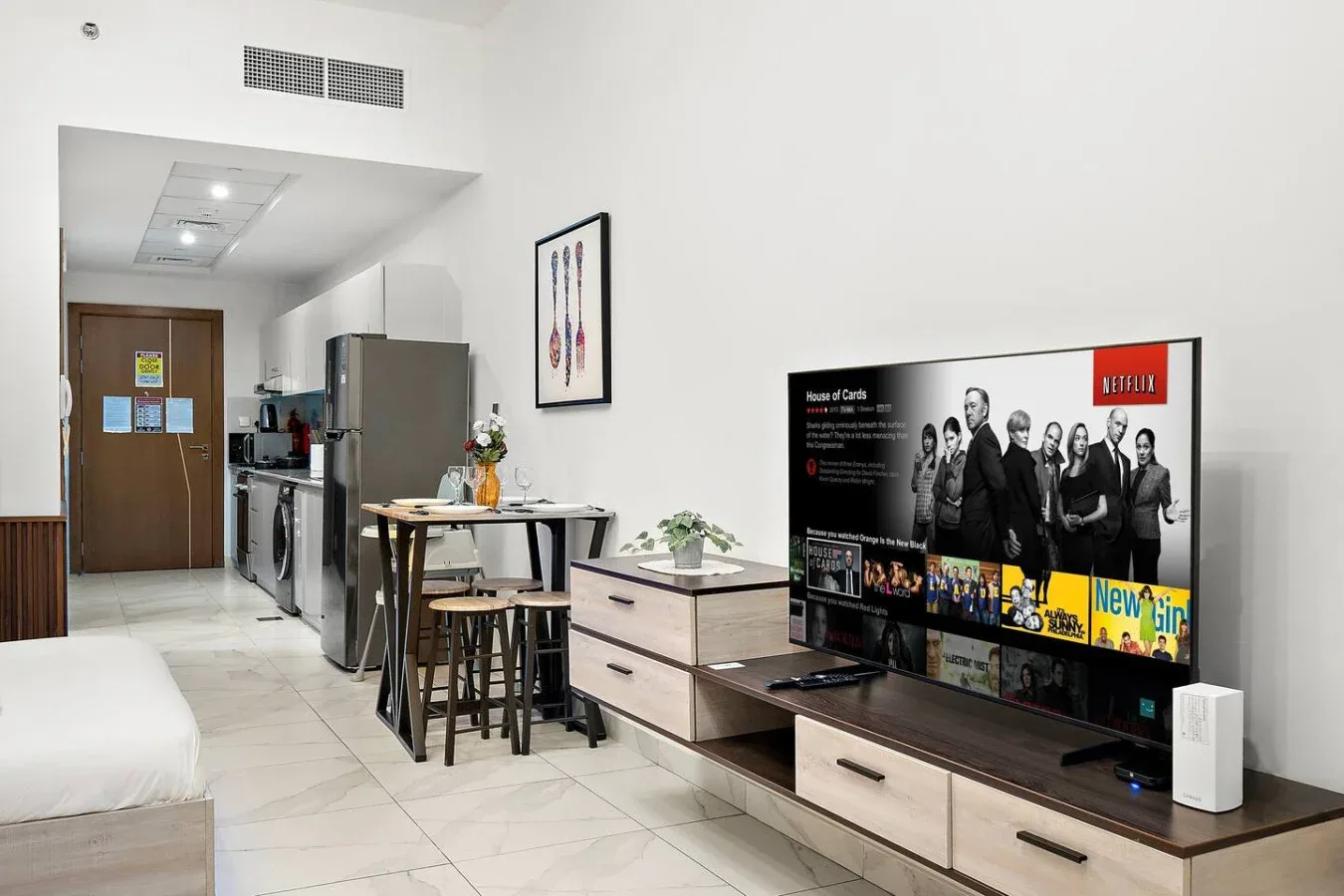 Un dormitorio y una pequeña cocina comparten un espacio abierto, con una mesa junto al televisor.