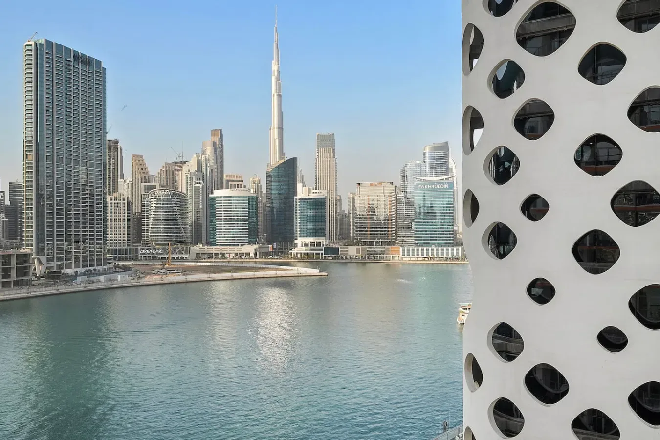 A waterfront city skyline rises across the water, with a distinctive patterned facade in the foreground.