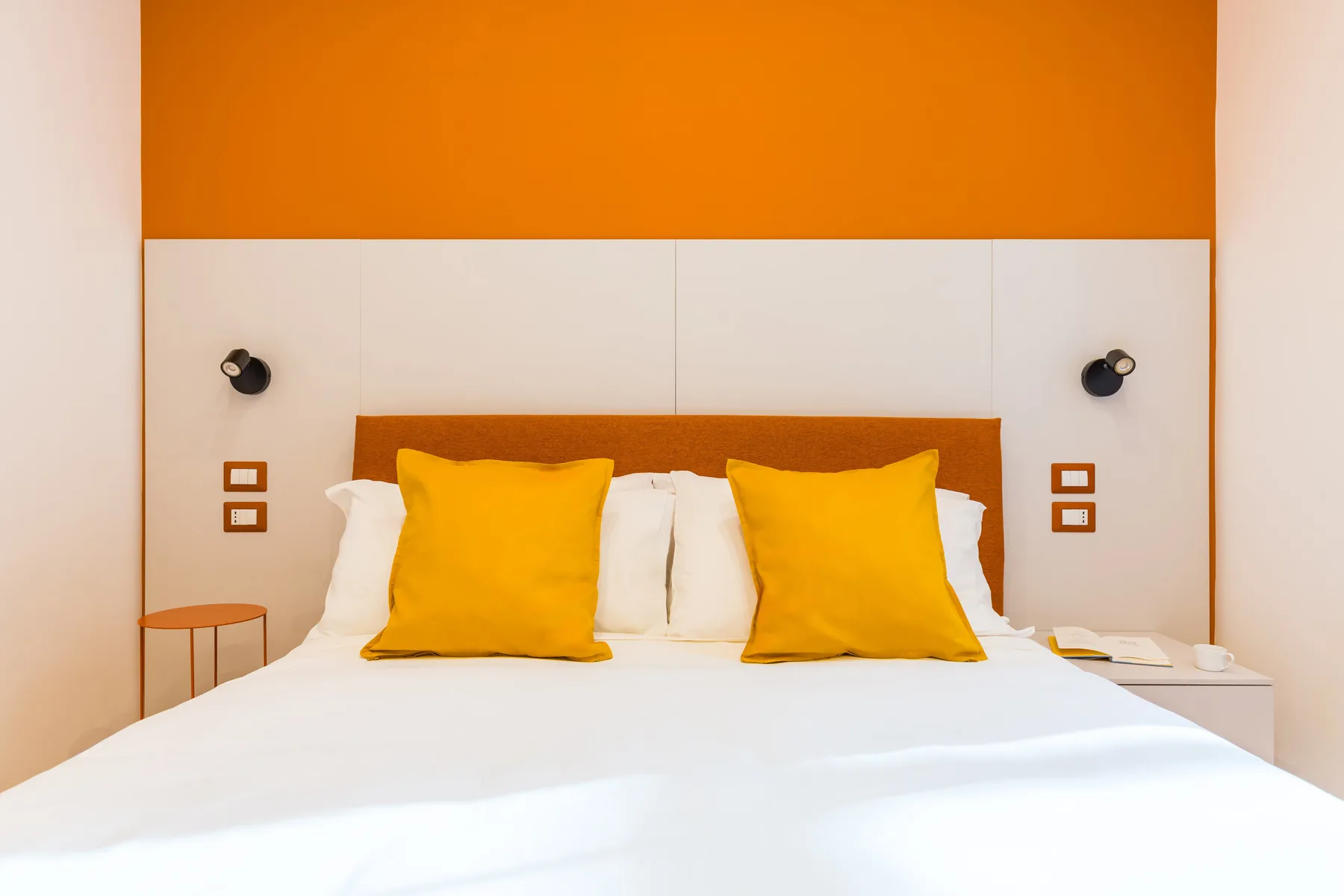 A bedroom with a white bed and bright yellow pillows against an orange wall.