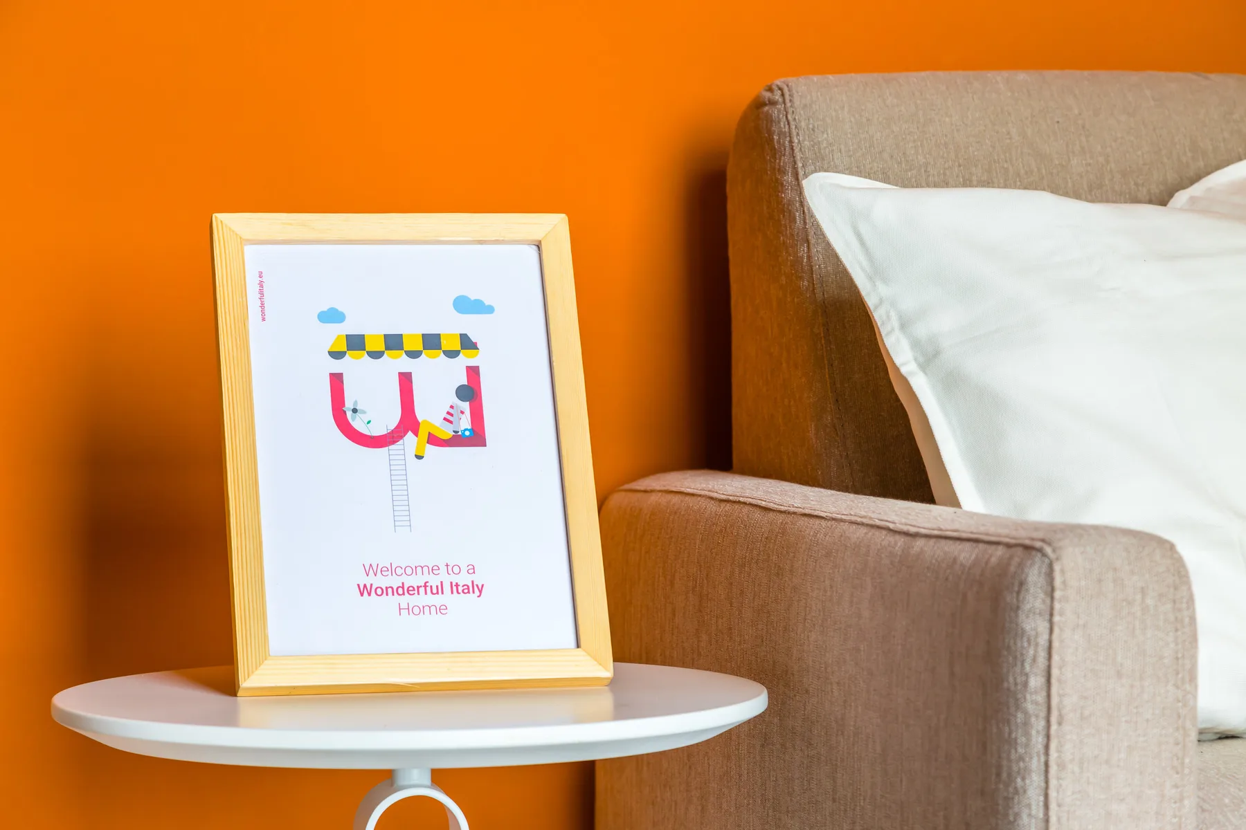 A framed print sits on a round side table beside a bed against an orange wall.