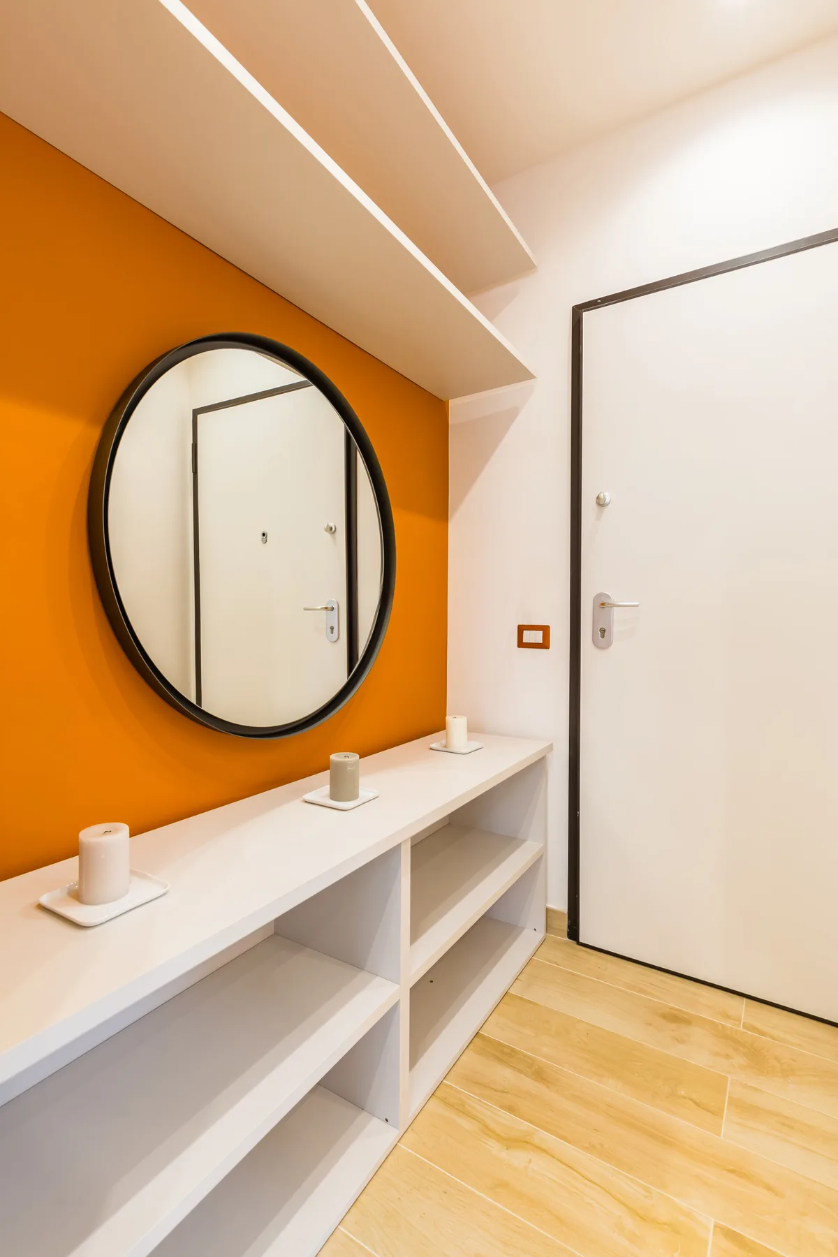 An entryway with an orange wall, round mirror, and white shelving.