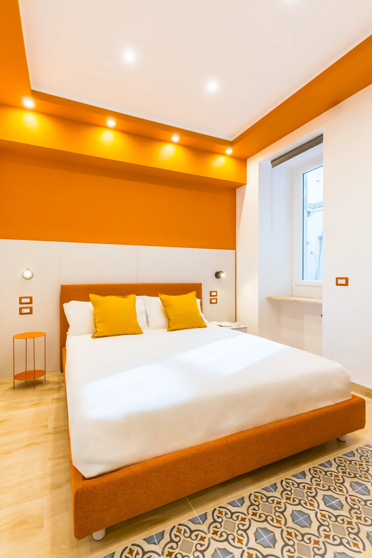 A bedroom with a white bed, orange accents, and a patterned rug.