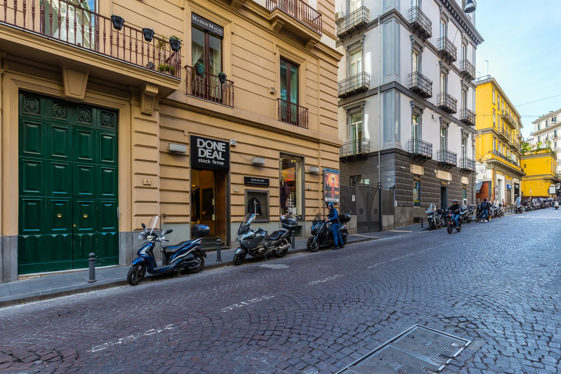 A cobblestone city street lined with multistory buildings and parked scooters.