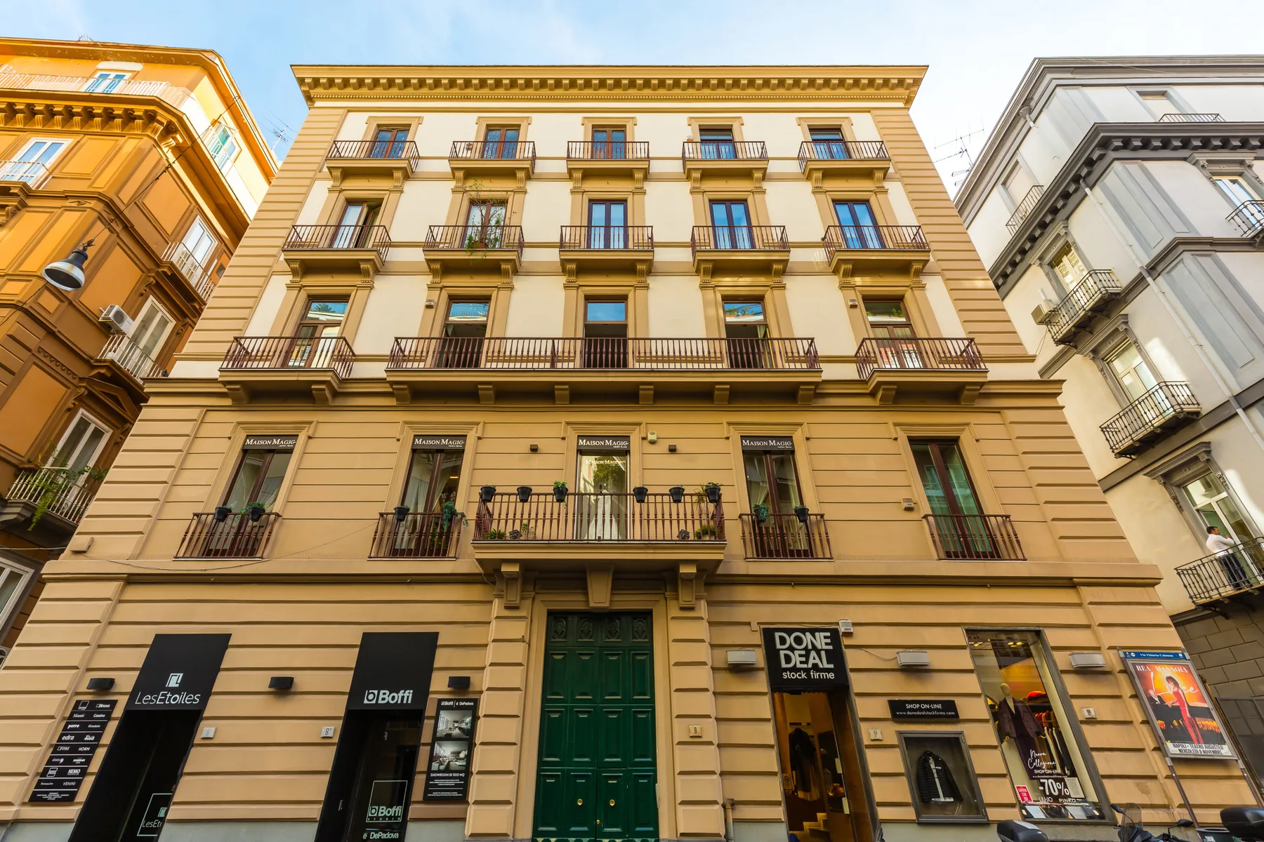 A tan multi-story building with rows of balconies and storefronts at street level.