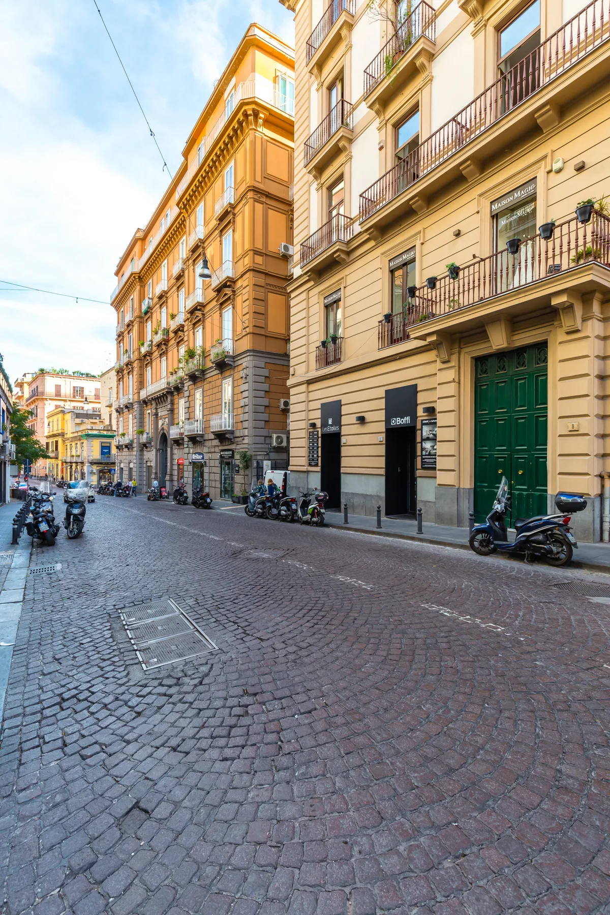 A cobblestone street runs between tall buildings, with scooters parked along the sidewalks.