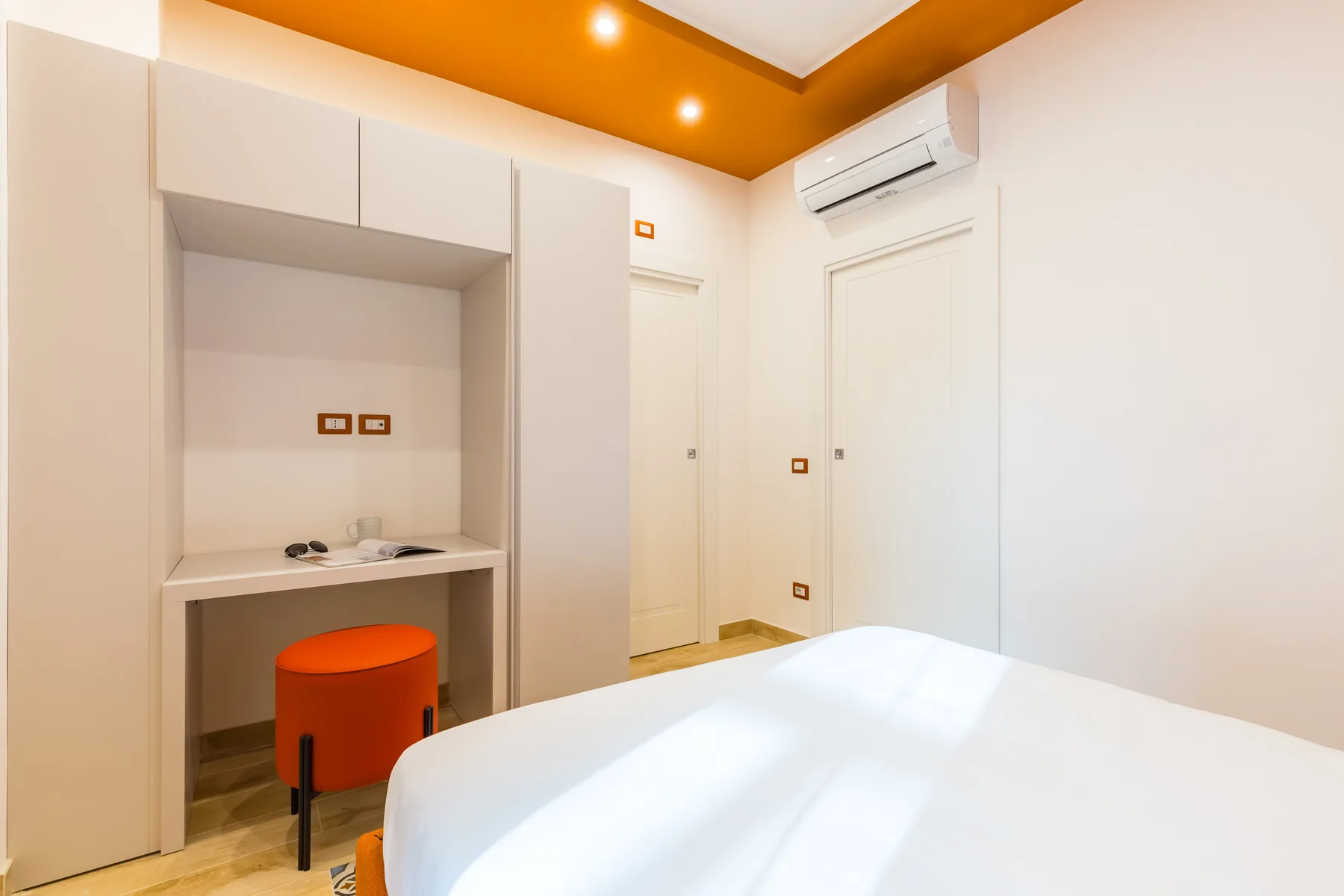 A bedroom with a white bed, built-in shelving, and a wall-mounted air conditioner.