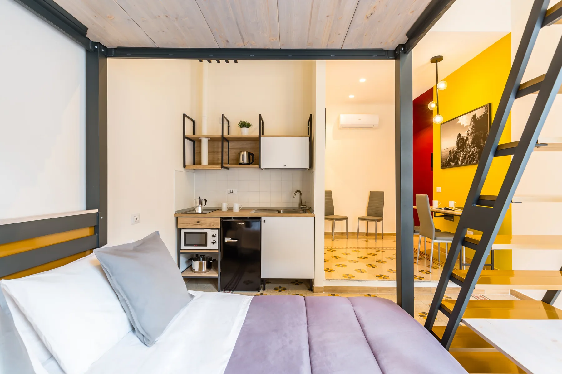 Un dormitorio compacto con cama elevada, cocina pequeña y zona de comedor.