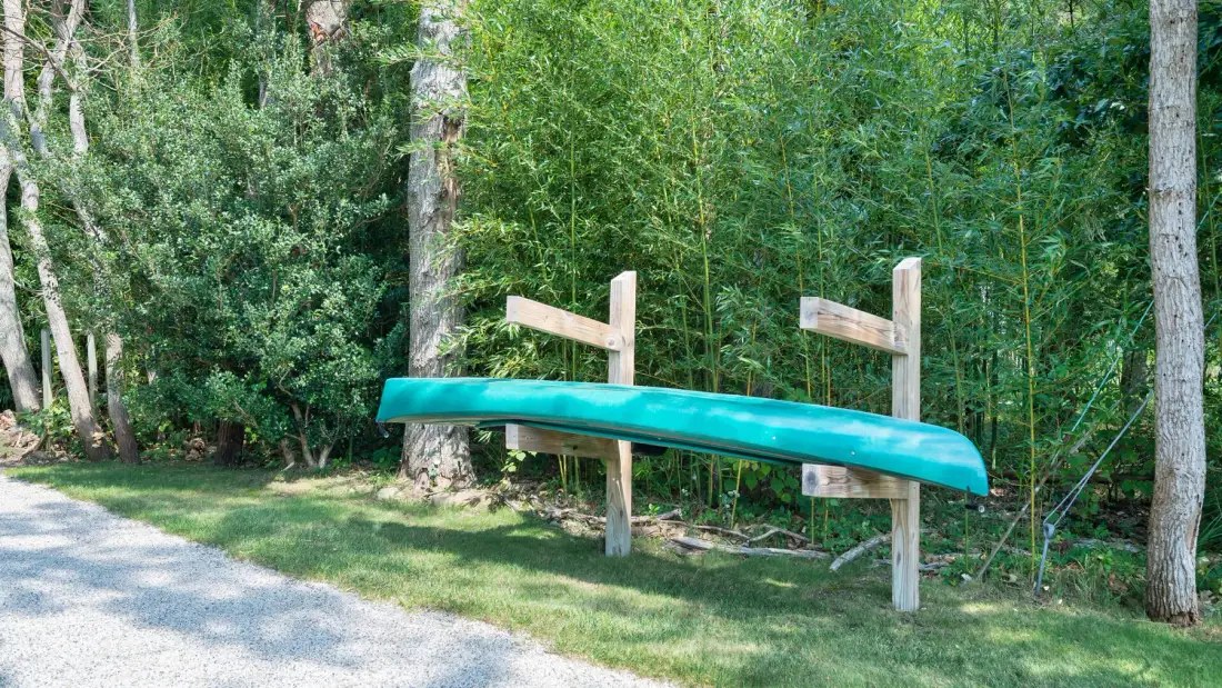 Una canoa verde descansa sobre soportes de madera junto a un camino de grava en una zona arbolada.